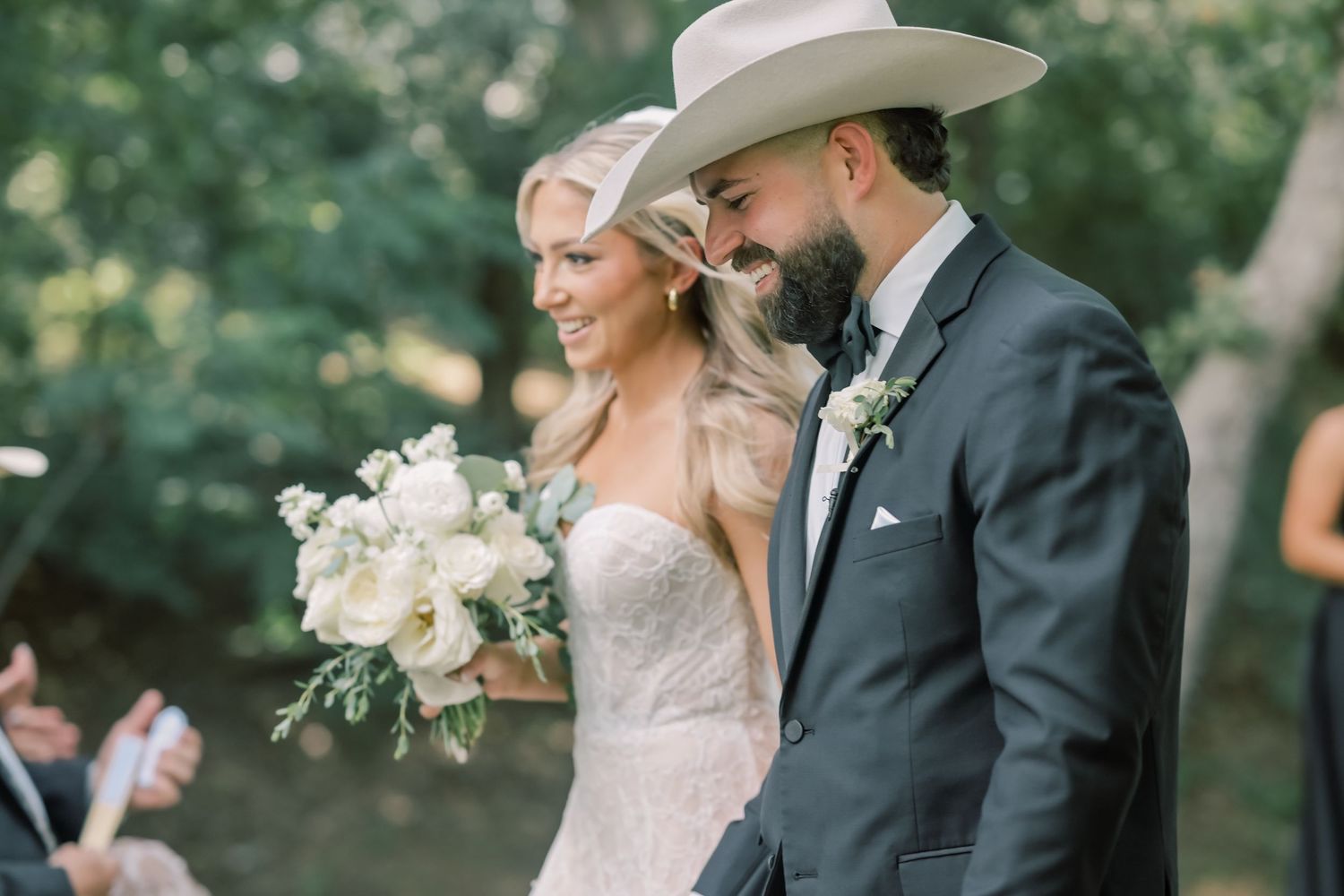 Smiling bride in lace gown with groom wearing a cowboy hat holding white floral bouquet.