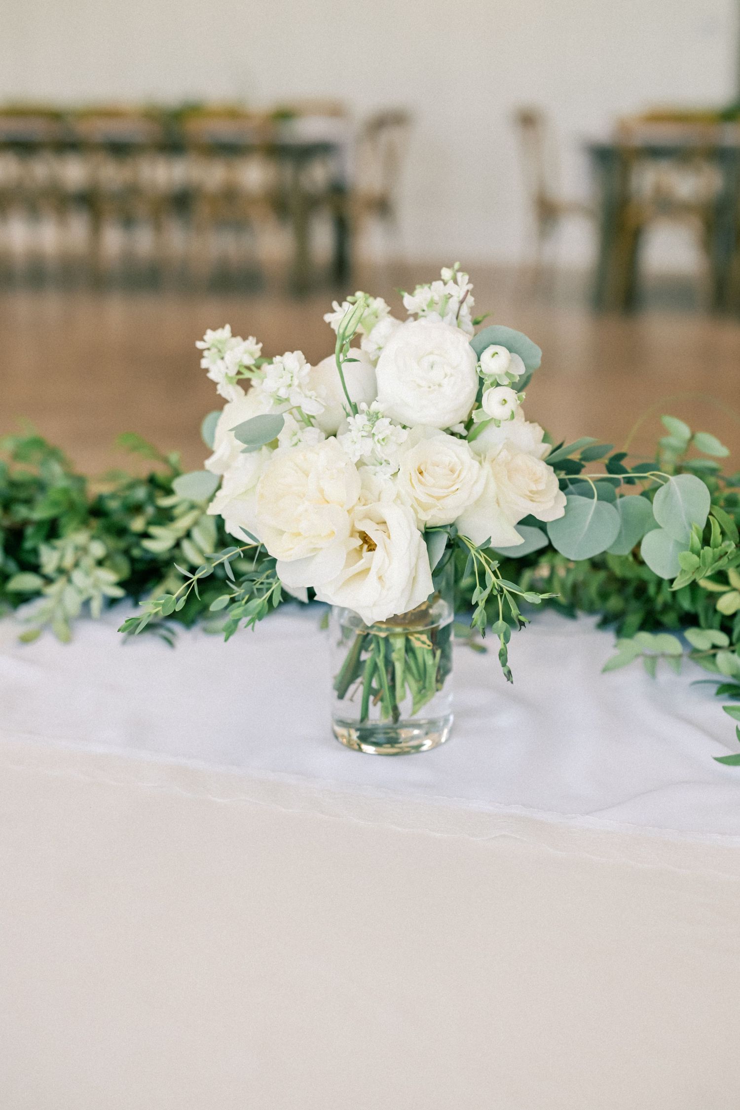 White roses and greenery centerpiece in glass vase on white tablecloth at wedding reception.