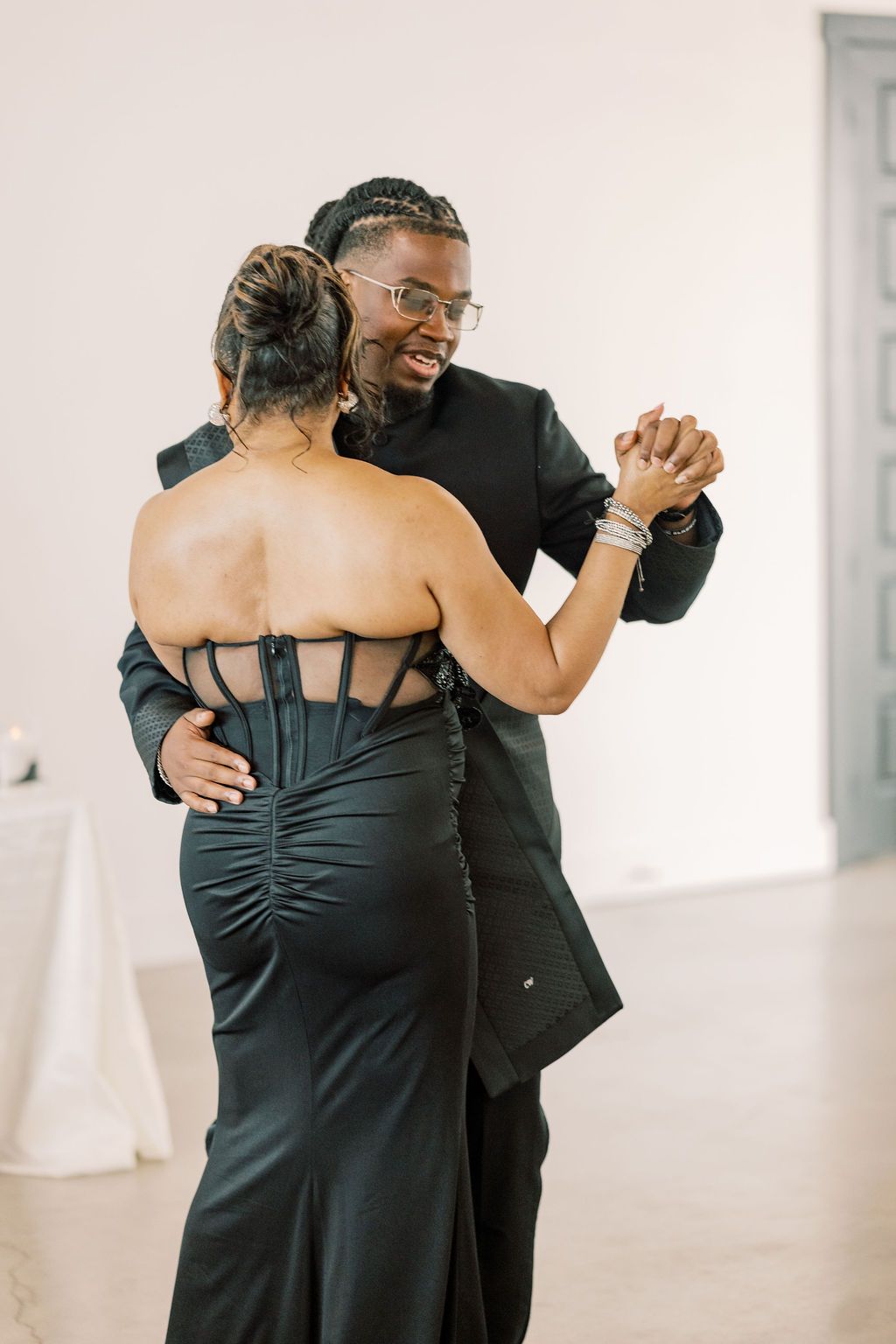 Couple dances closely at wedding reception, man in black suit smiling, woman in elegant backless black gown.