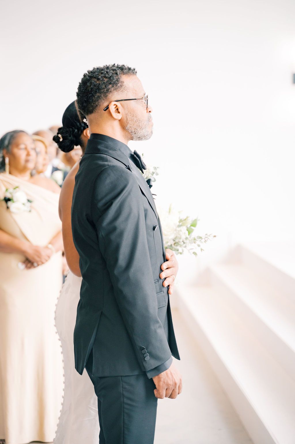 Groom in dark suit stands at altar holding greenery boutonniere during wedding ceremony.