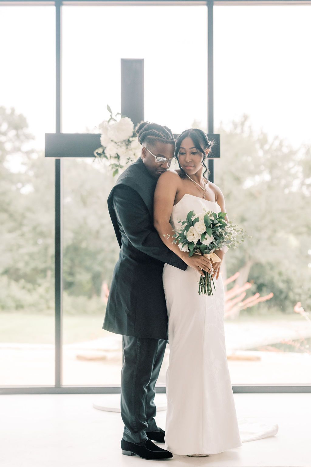 Bride and groom embrace before a cross at their elegant wedding ceremony, bride holding white floral bouquet.