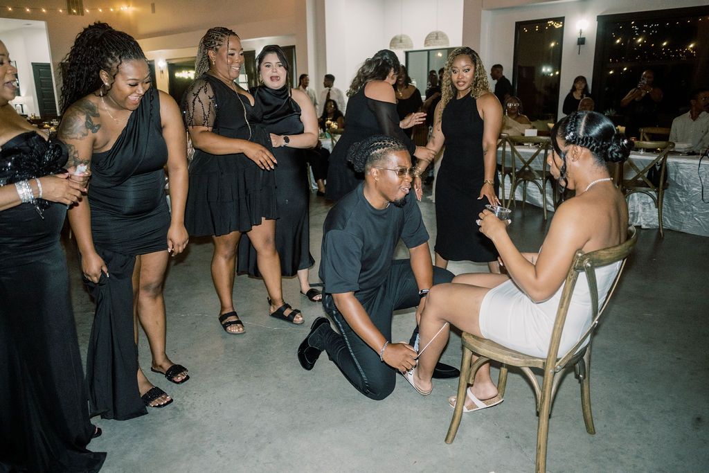 Wedding guests in black dresses watch as groom kneels to put shoes on bride seated in wooden chair.