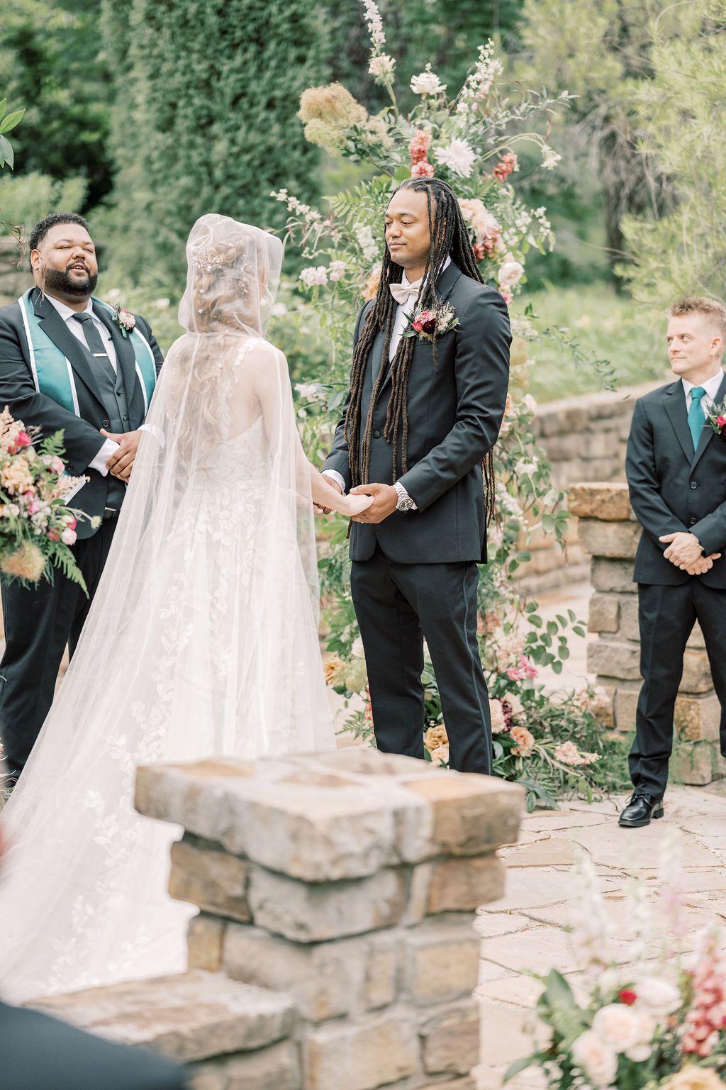 Groom in black suit holds bride's hands at outdoor wedding ceremony with floral arch and wedding party.