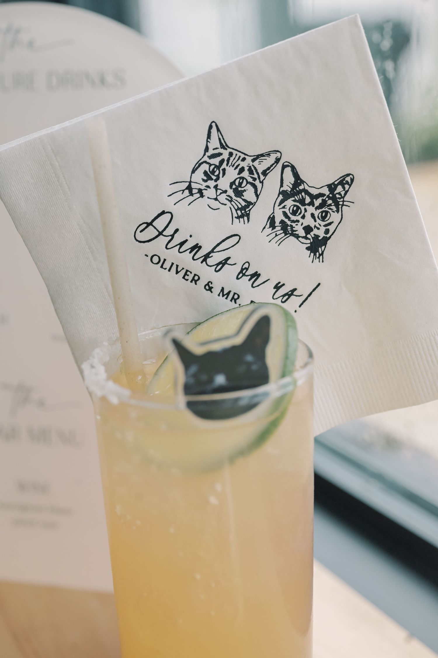 A yellow cocktail with a cat face stirrer and custom napkin reading Drinks on us Oliver and Mr at a wedding bar.