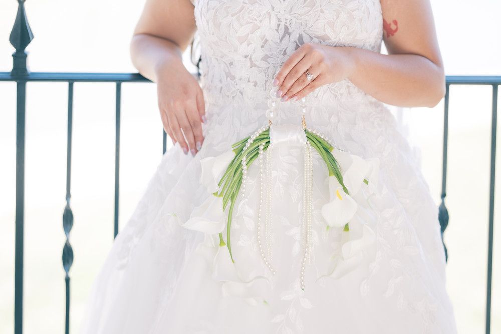 Midsection of bride in lace gown holding white calla lily bouquet against metal railing.