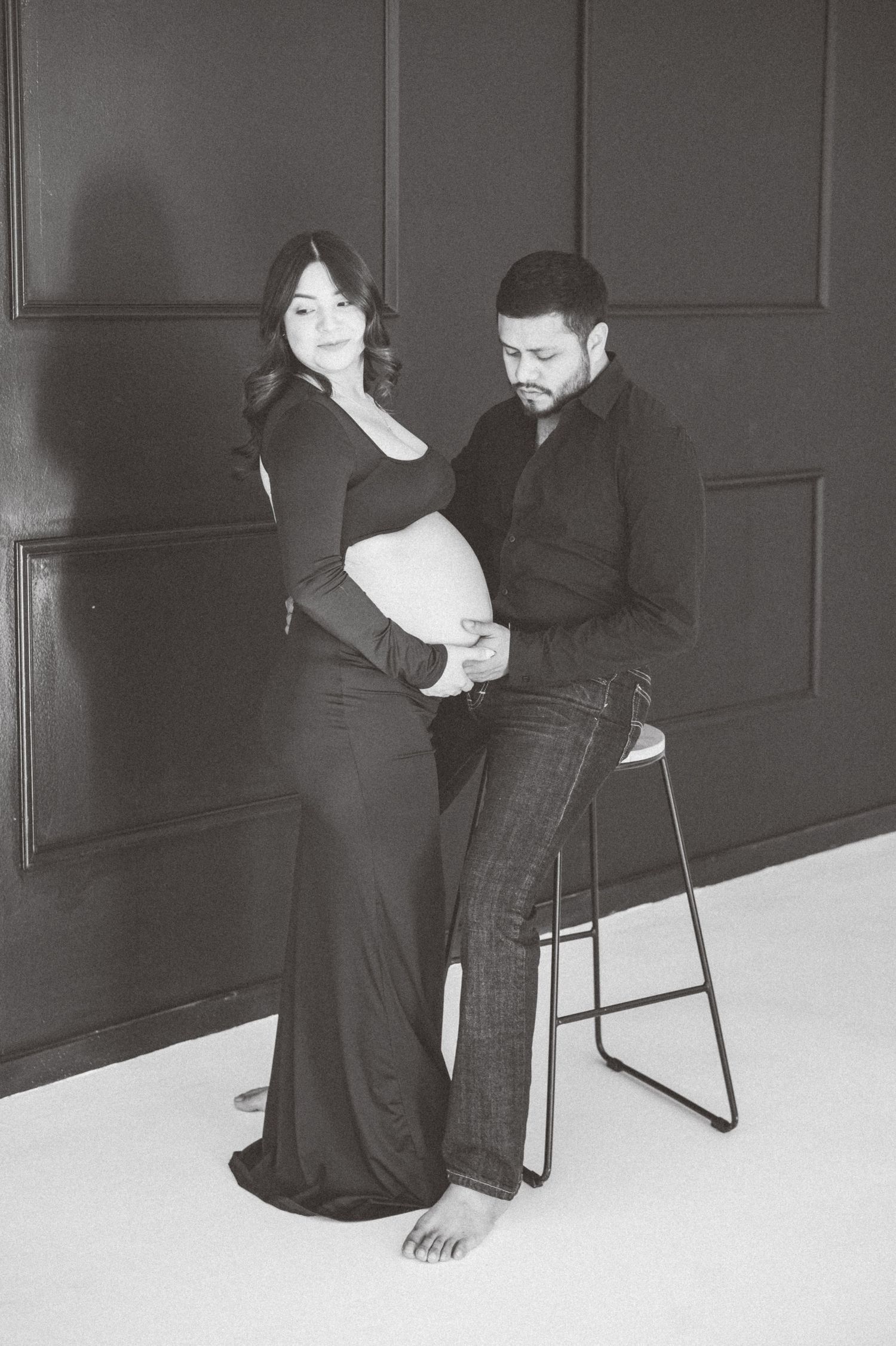 Black and white maternity photo of a couple, man seated on stool touching pregnant womans belly in studio.