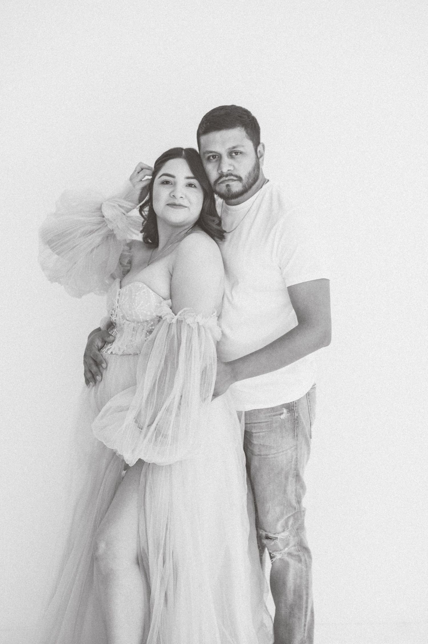 Black and white photo of a couple posing together, woman in elegant tulle gown, man in casual attire, studio setting.