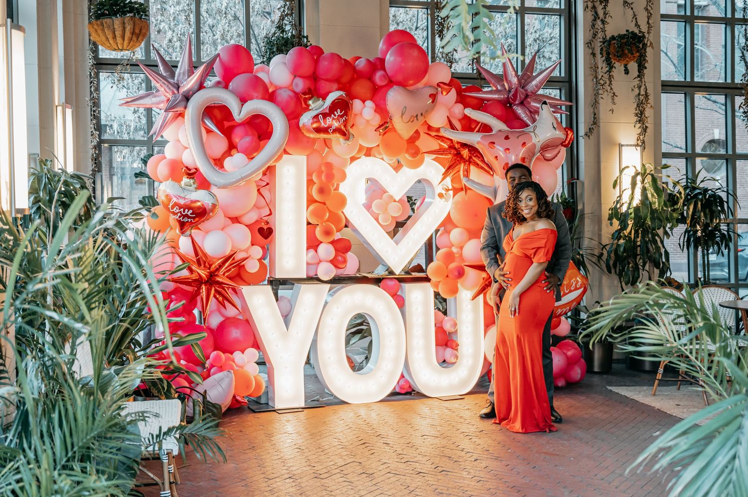 A woman in a red gown poses beside a large illuminated I Love You sign surrounded by red and pink balloon decor.