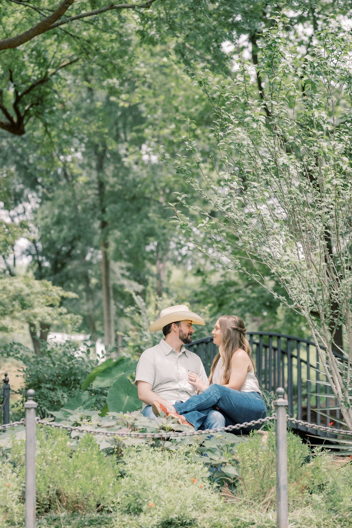 Couple sitting together on a blanket in a lush green garden, man wearing cowboy hat, sharing a romantic moment outdoors.
