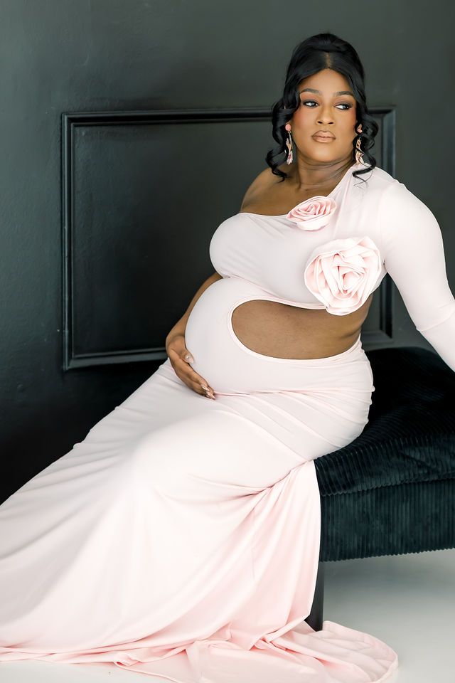 Expectant mother in pink one-shoulder cutout dress with rose applique posing on dark velvet bench.