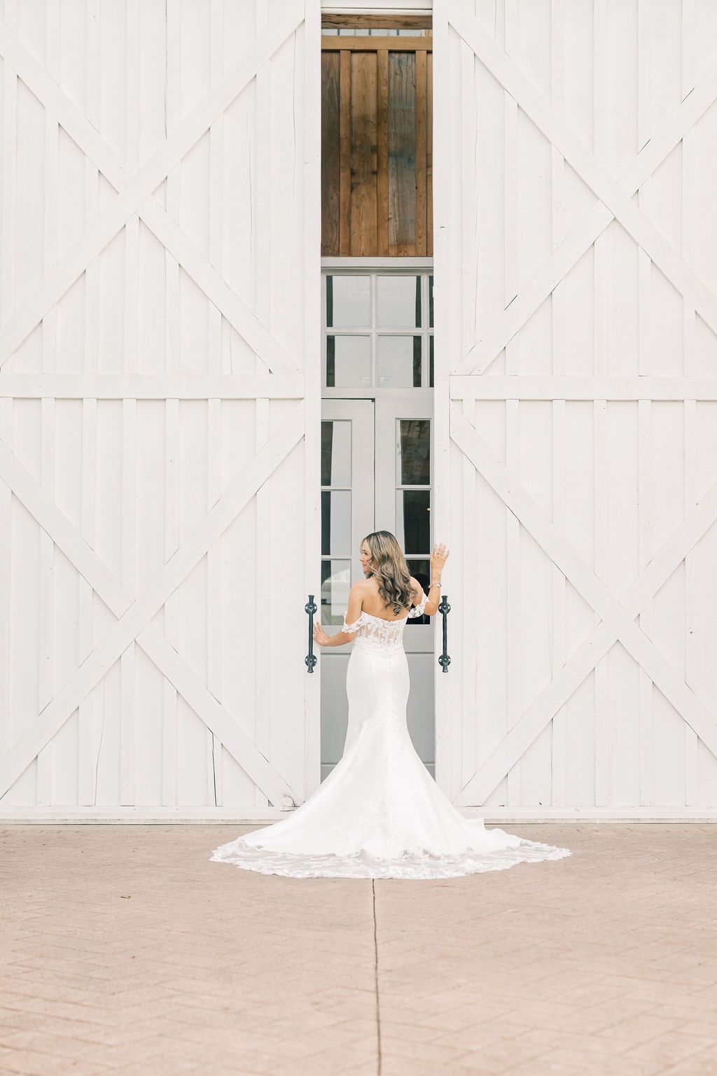 Bride in elegant mermaid wedding gown with lace train stands before white barn doors at rustic wedding venue.
