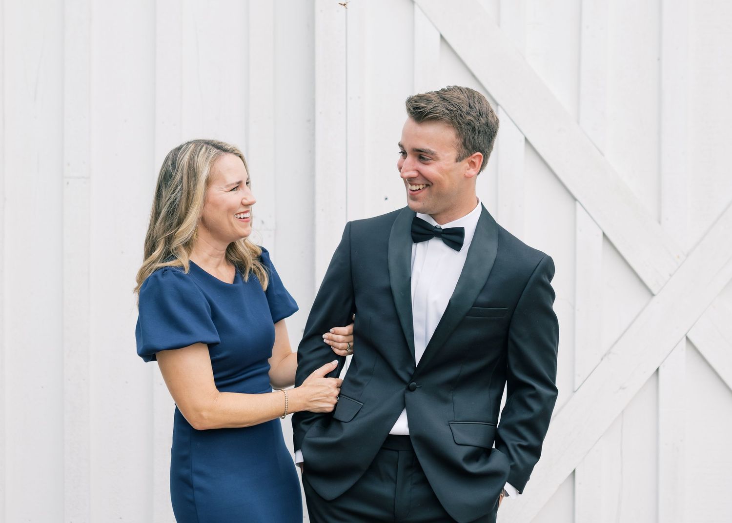Groom in black tuxedo laughs with woman in navy dress outside white barn wedding venue.