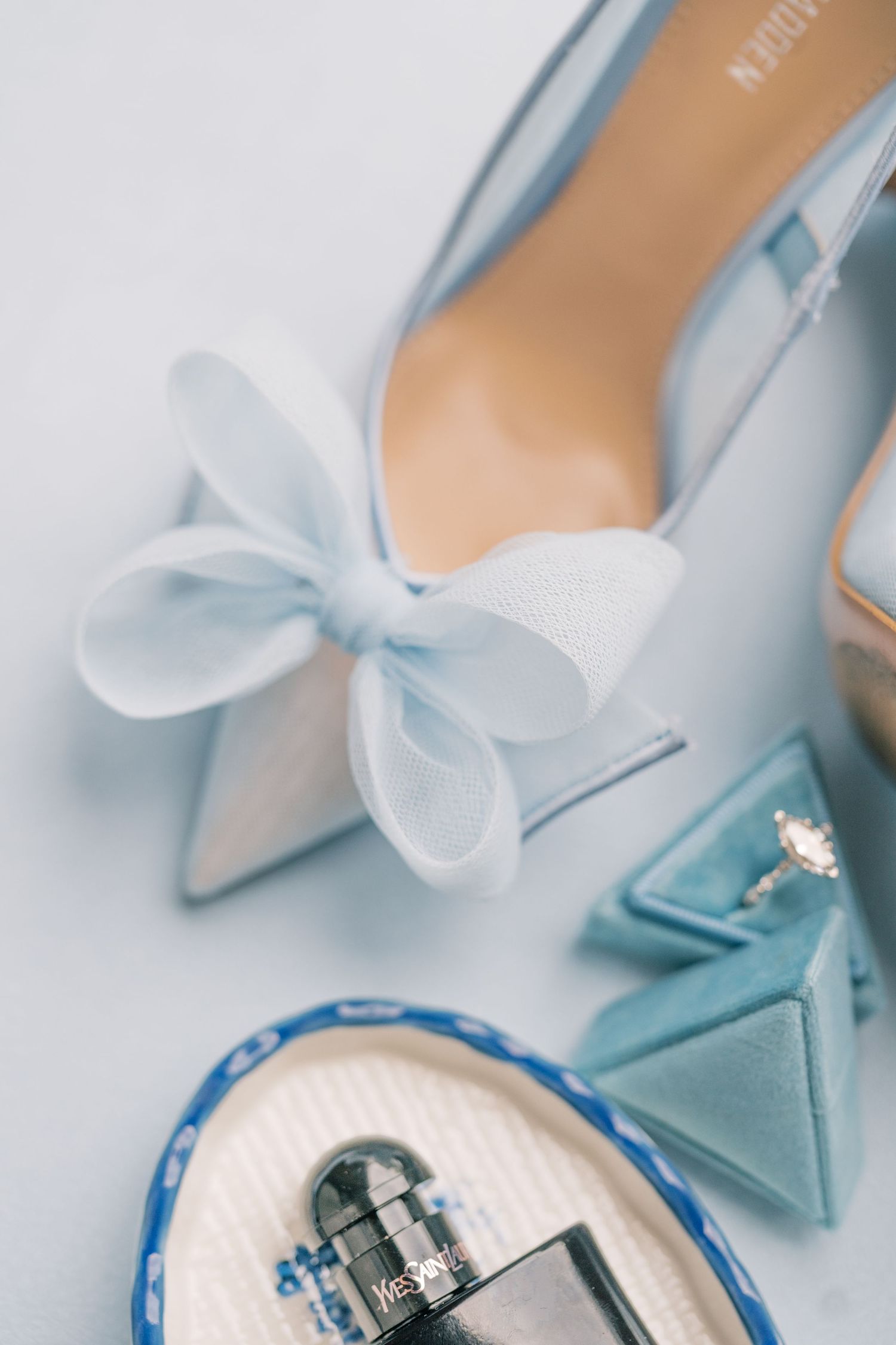 Light blue bridal heels with bow detail beside a velvet ring box and perfume bottle on a soft blue background.