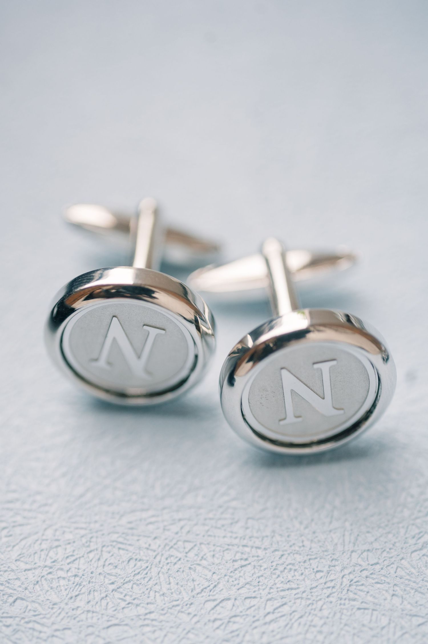 Silver cufflinks engraved with the letter N on a light blue background.