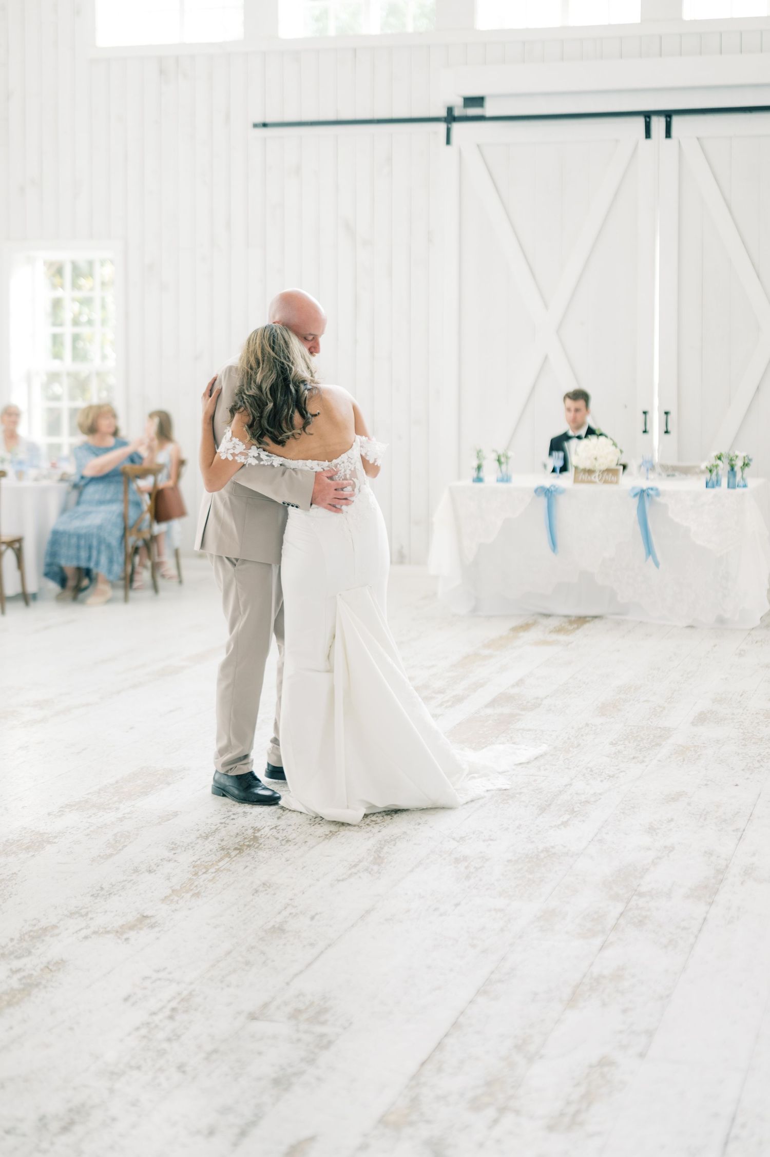 Bride and father sharing emotional first dance in white barn reception venue with guests seated.