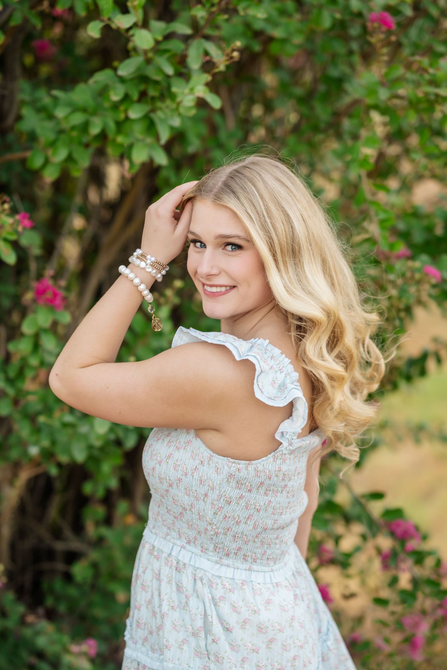 Smiling blonde teen girl in white floral dress poses outdoors near blooming pink flowers and green foliage.