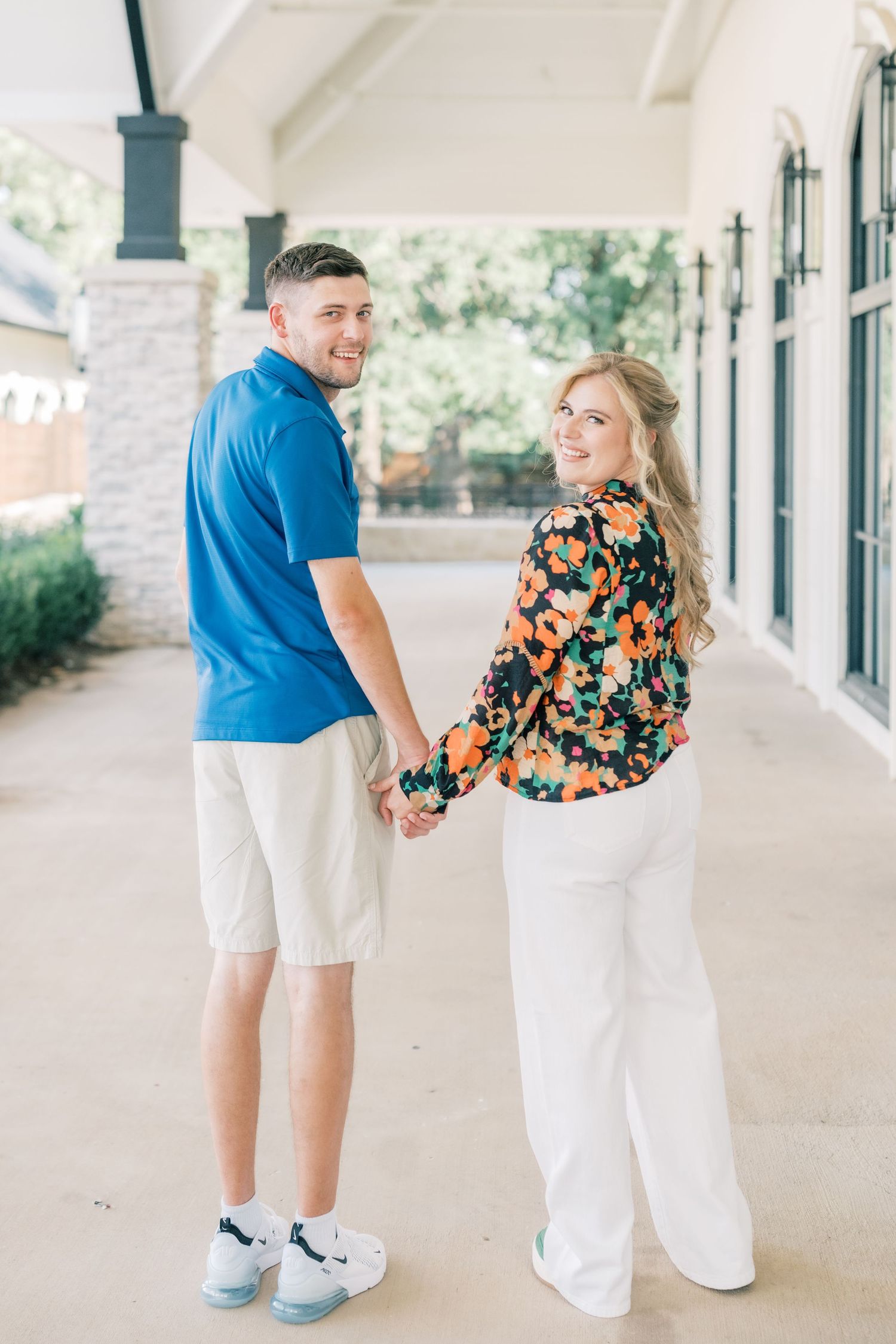 Smiling couple holding hands and looking back at camera in covered walkway, dressed in coordinating outfits.