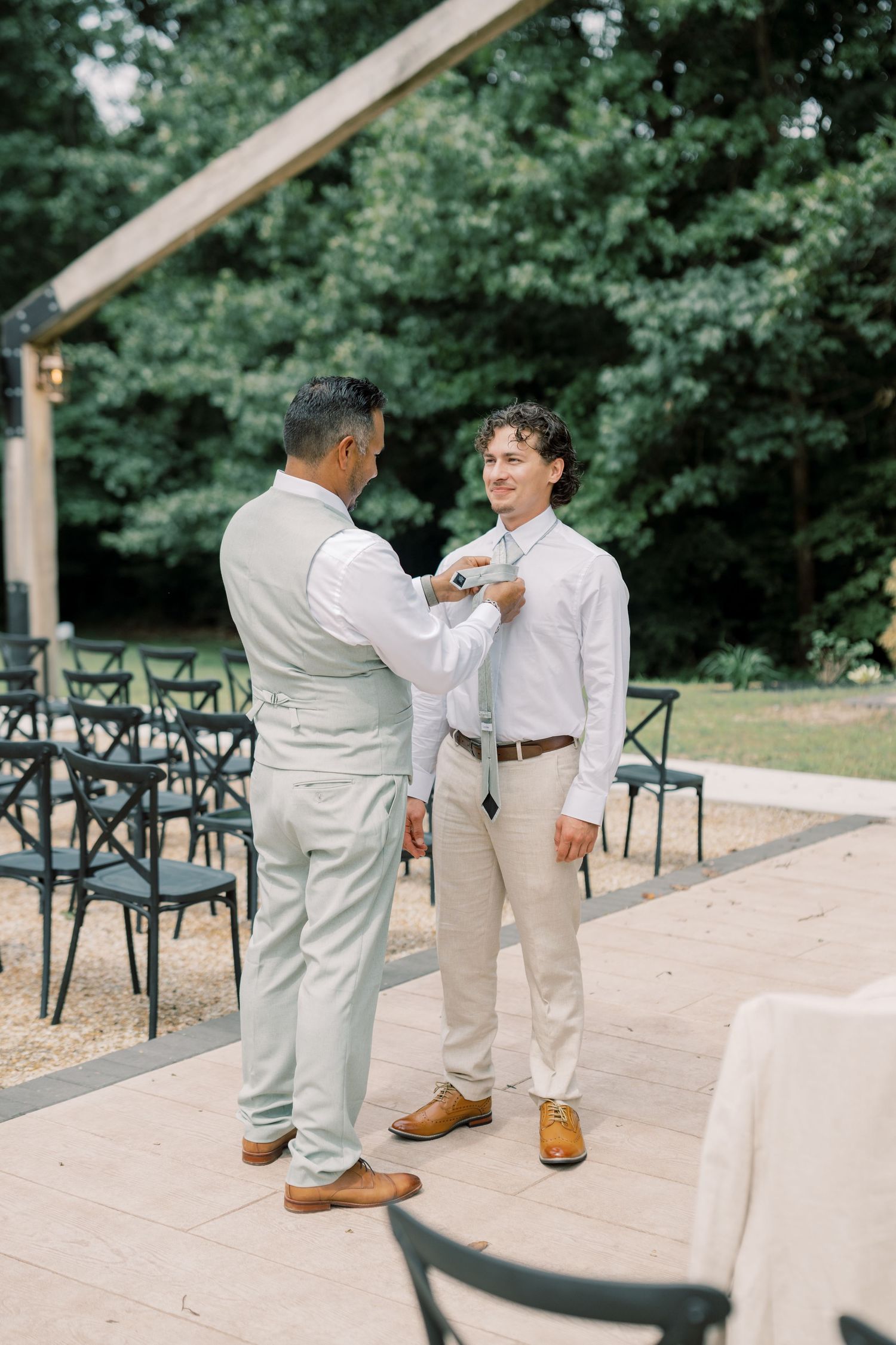 Two men in light suits pin boutonnieres outdoors at a wedding ceremony with chairs and greenery in background.