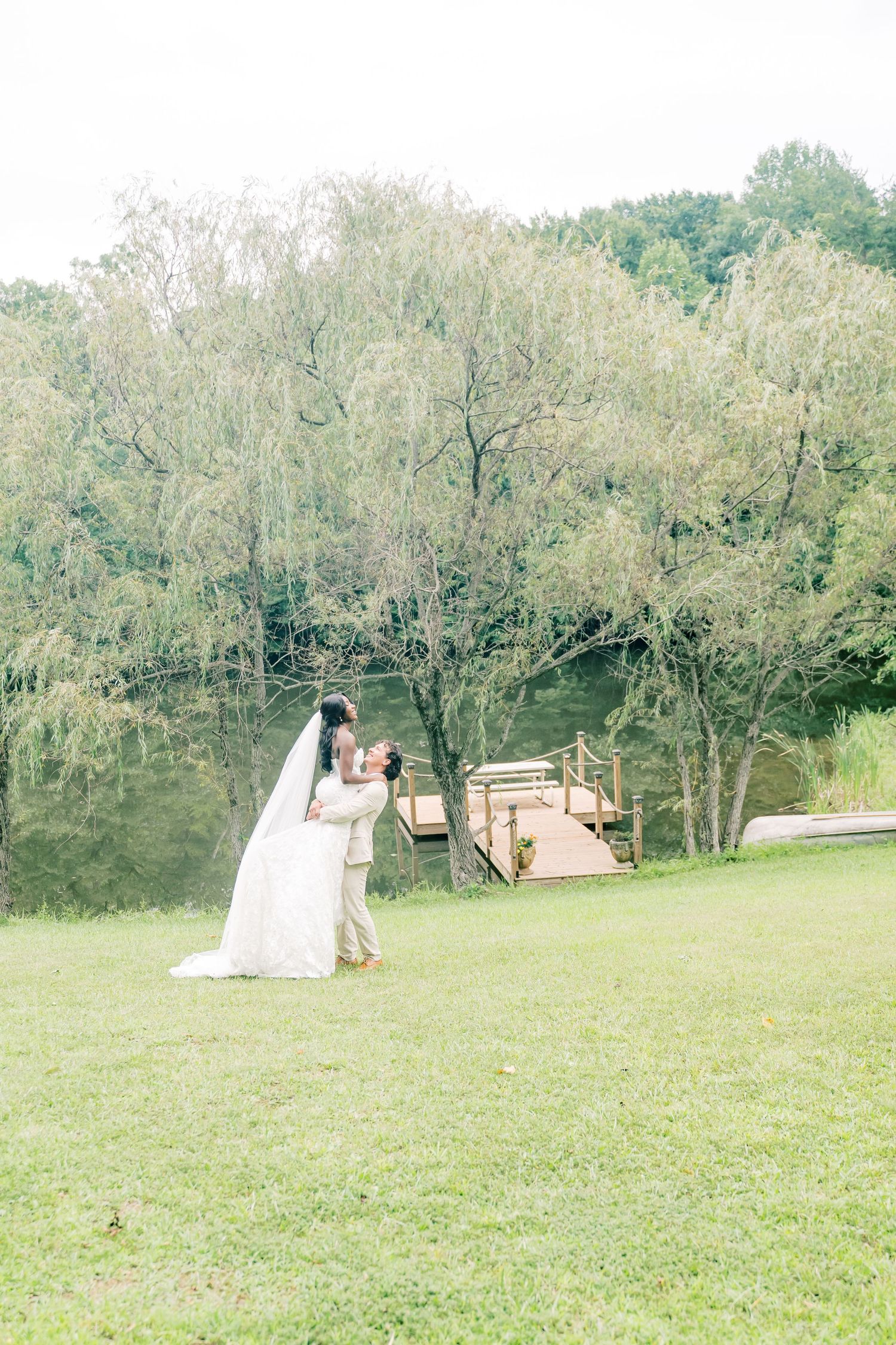 Bride and groom share romantic embrace on green lawn near wooden dock and weeping willow trees.