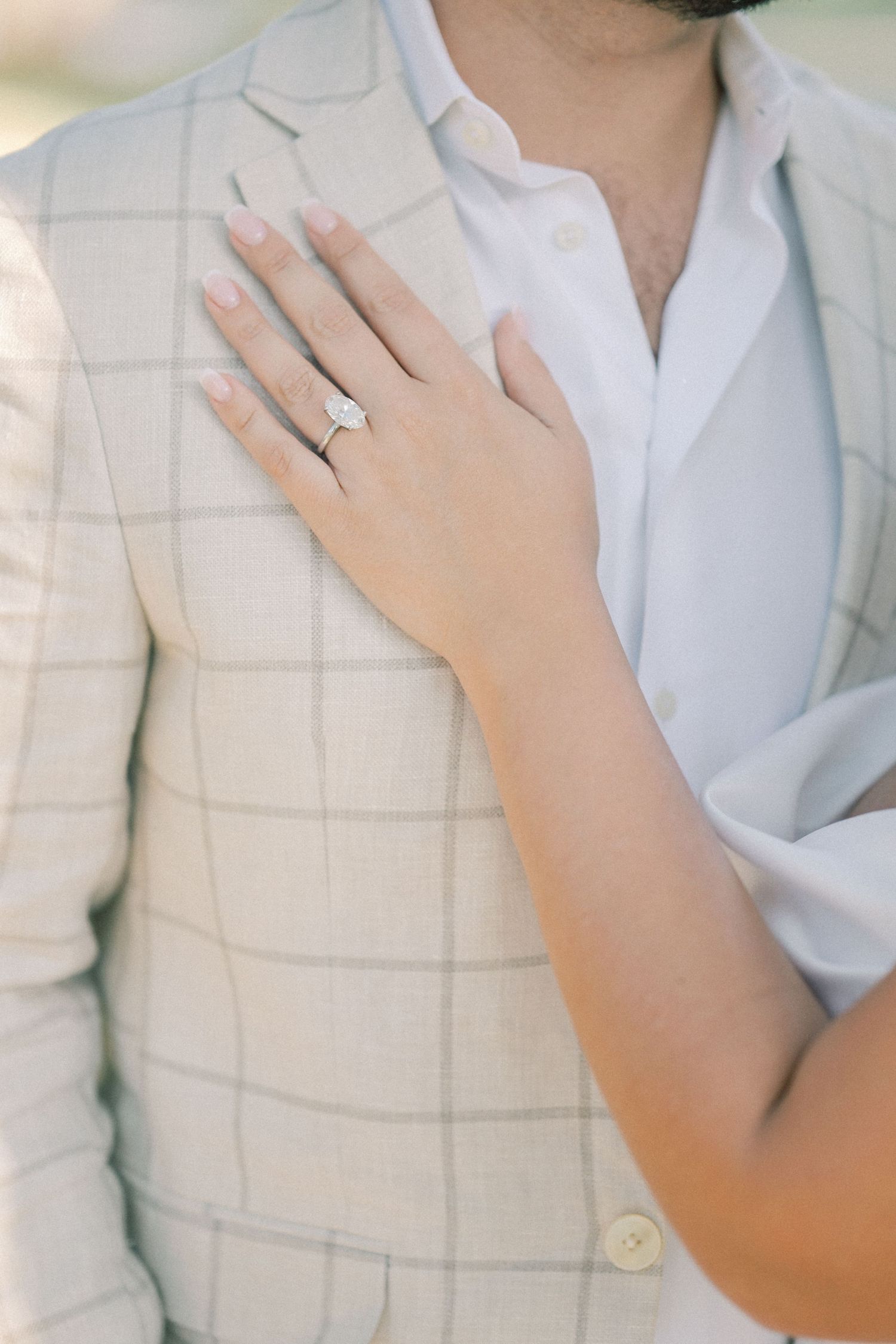 Close-up of a woman's hand with a sparkling diamond engagement ring resting on a man's cream plaid blazer.