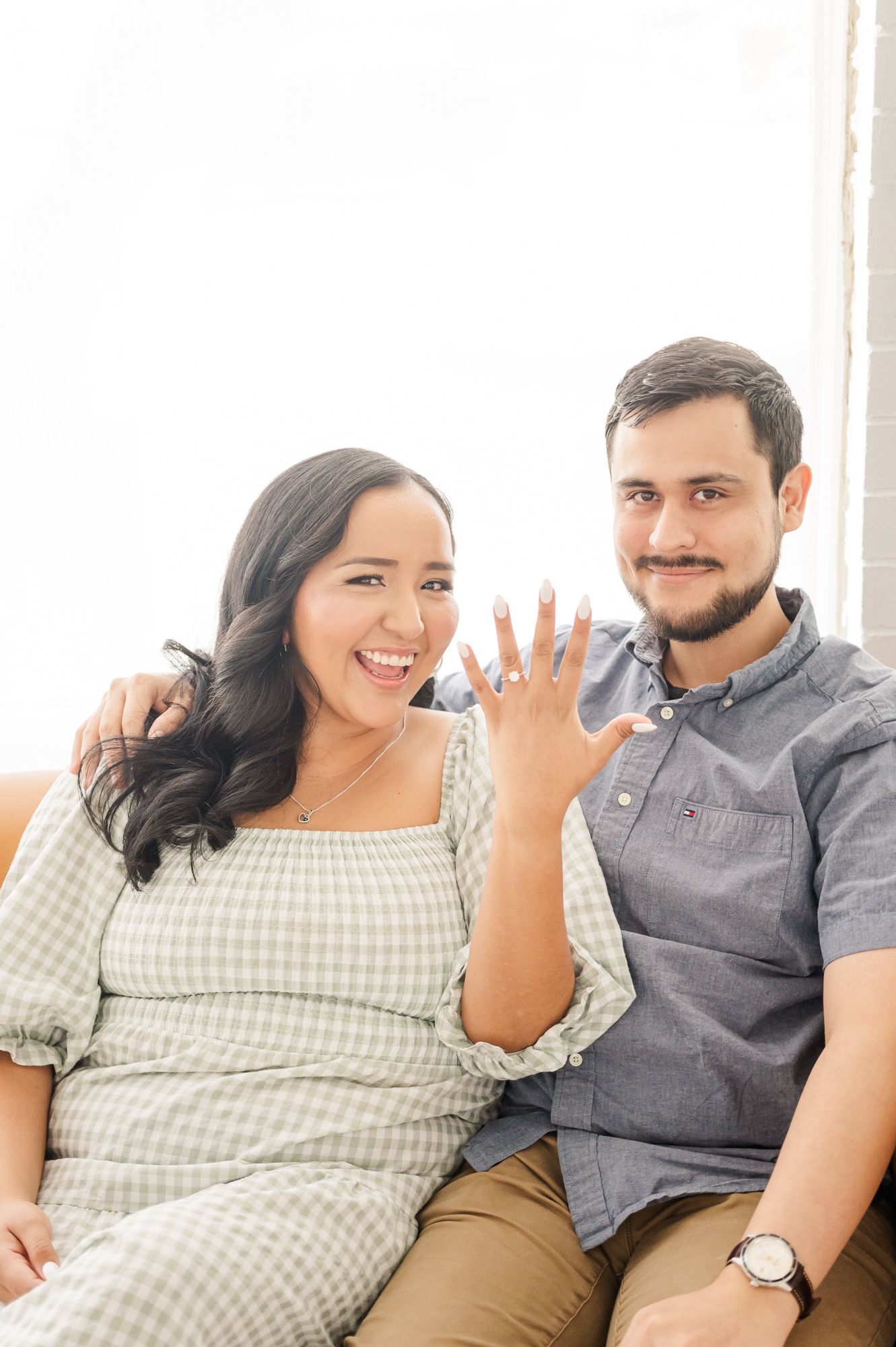 Newly engaged couple sitting together and showing off the ring with joyful expressions.