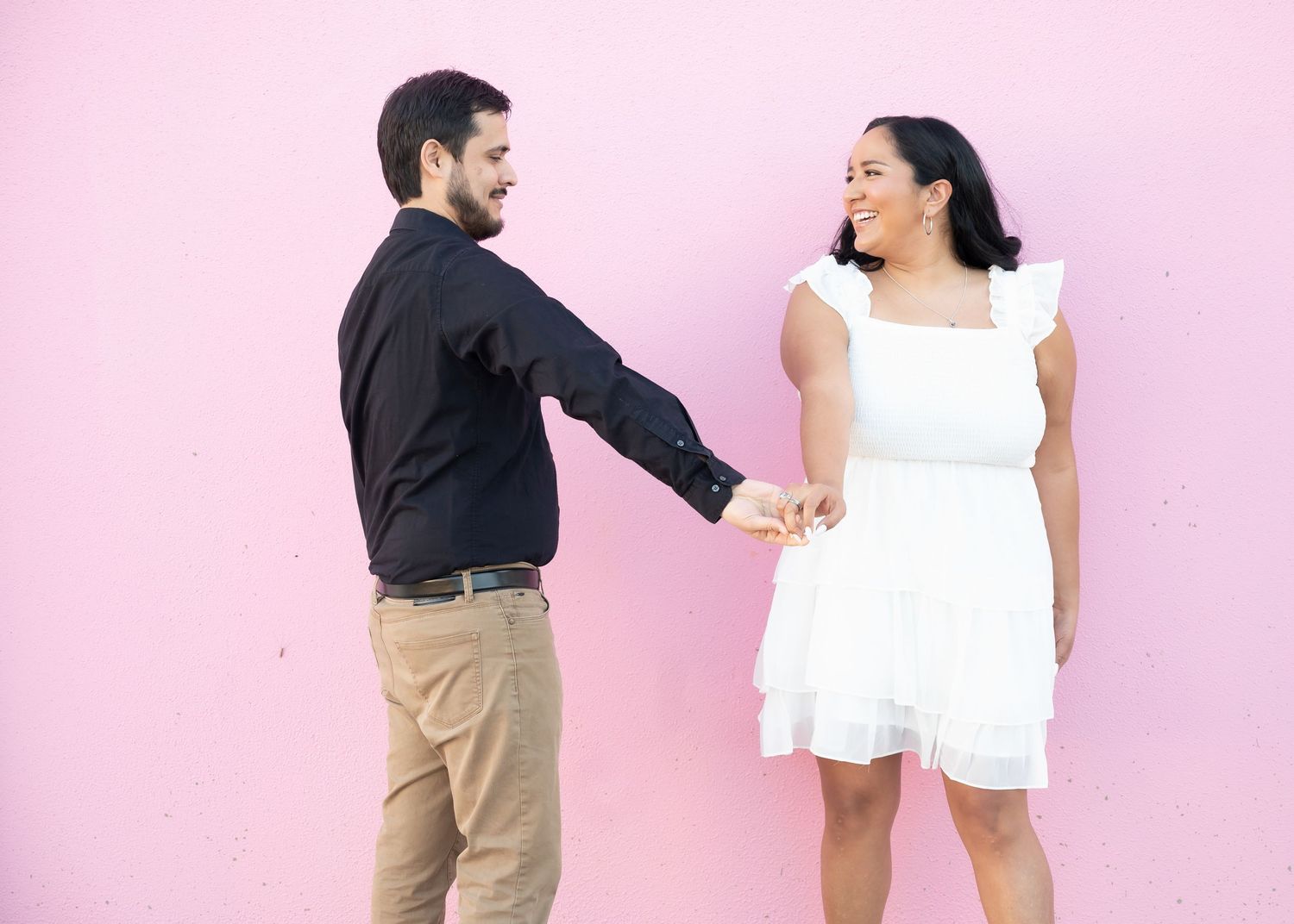 A couple in contrasting outfits poses playfully against a pink wall while holding hands.