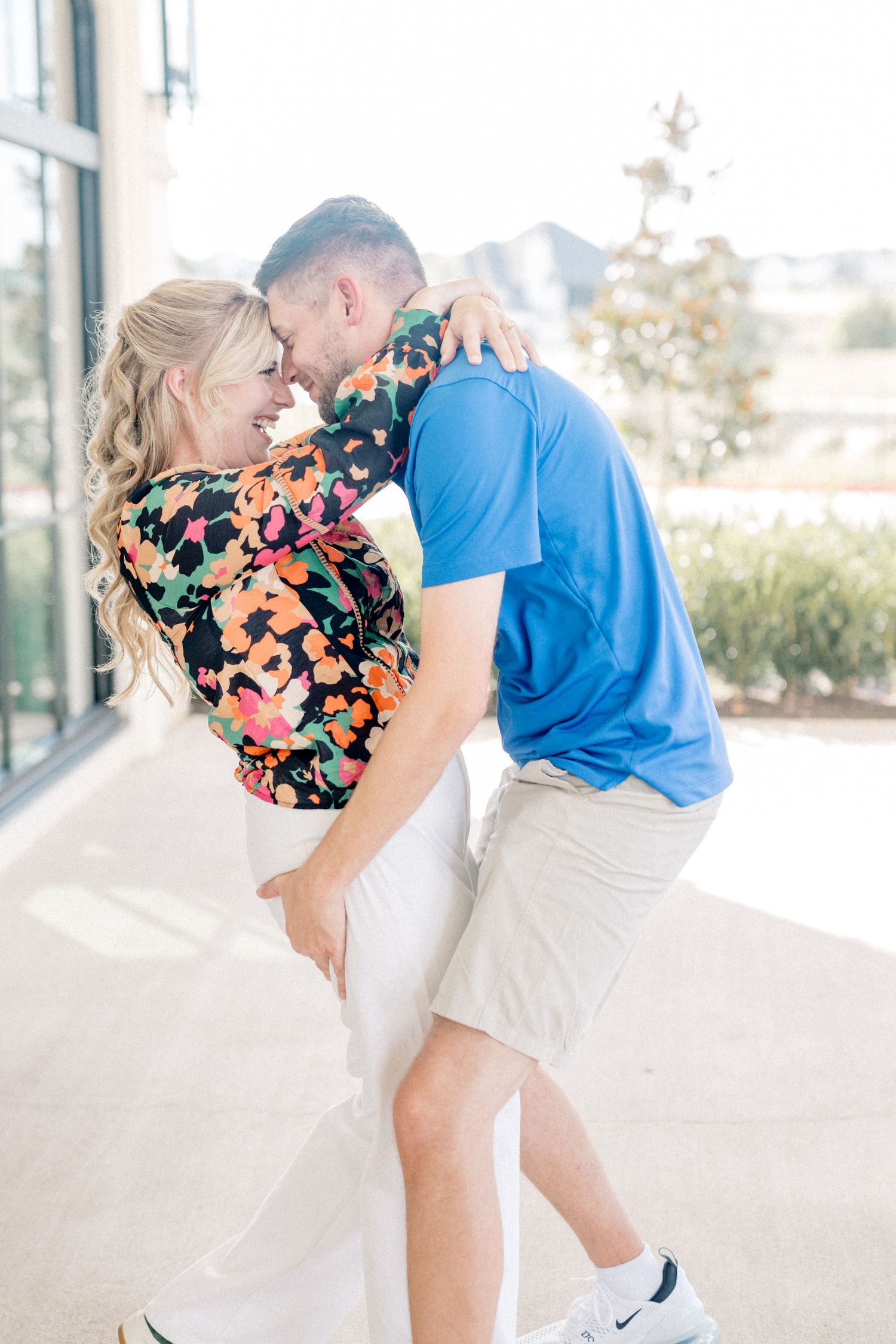 Couple embracing and laughing outdoors, woman in floral top and man in blue polo near building.