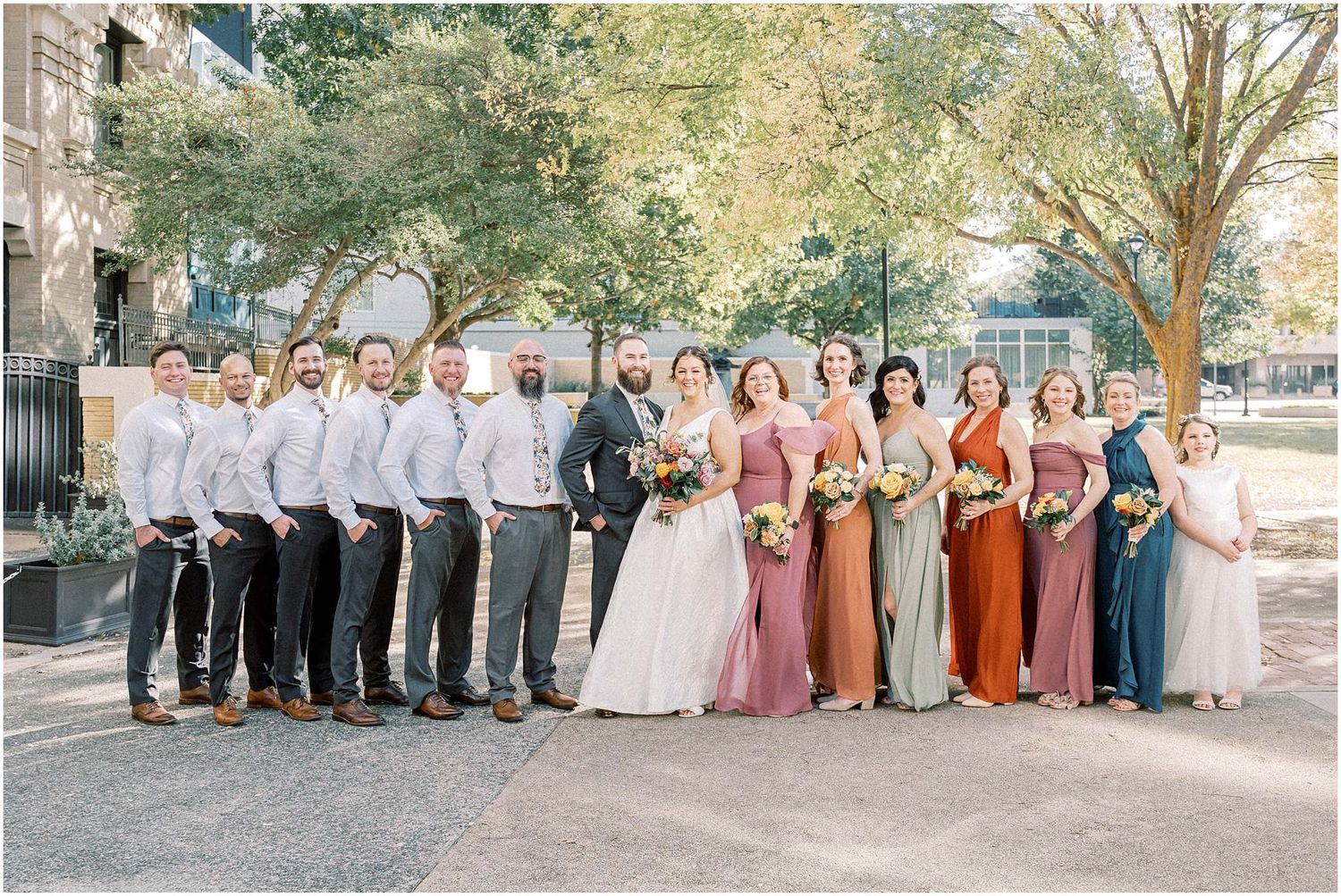 Wedding party in coordinated attire featuring bridesmaids in warm autumn tones and groomsmen in light gray suits.