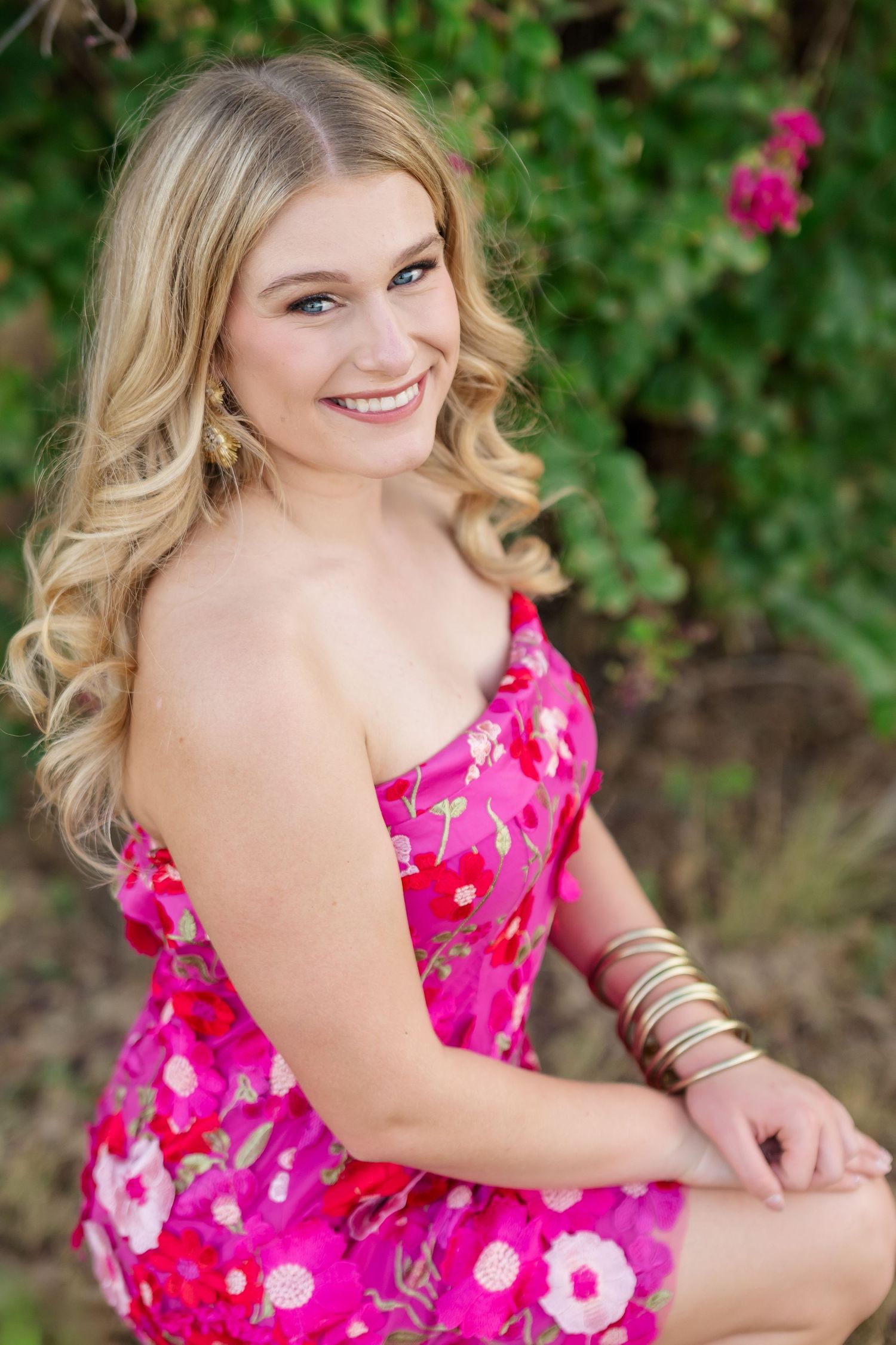 Smiling blonde woman in pink floral dress with gold bracelets, sitting outdoors near green foliage and pink flowers.