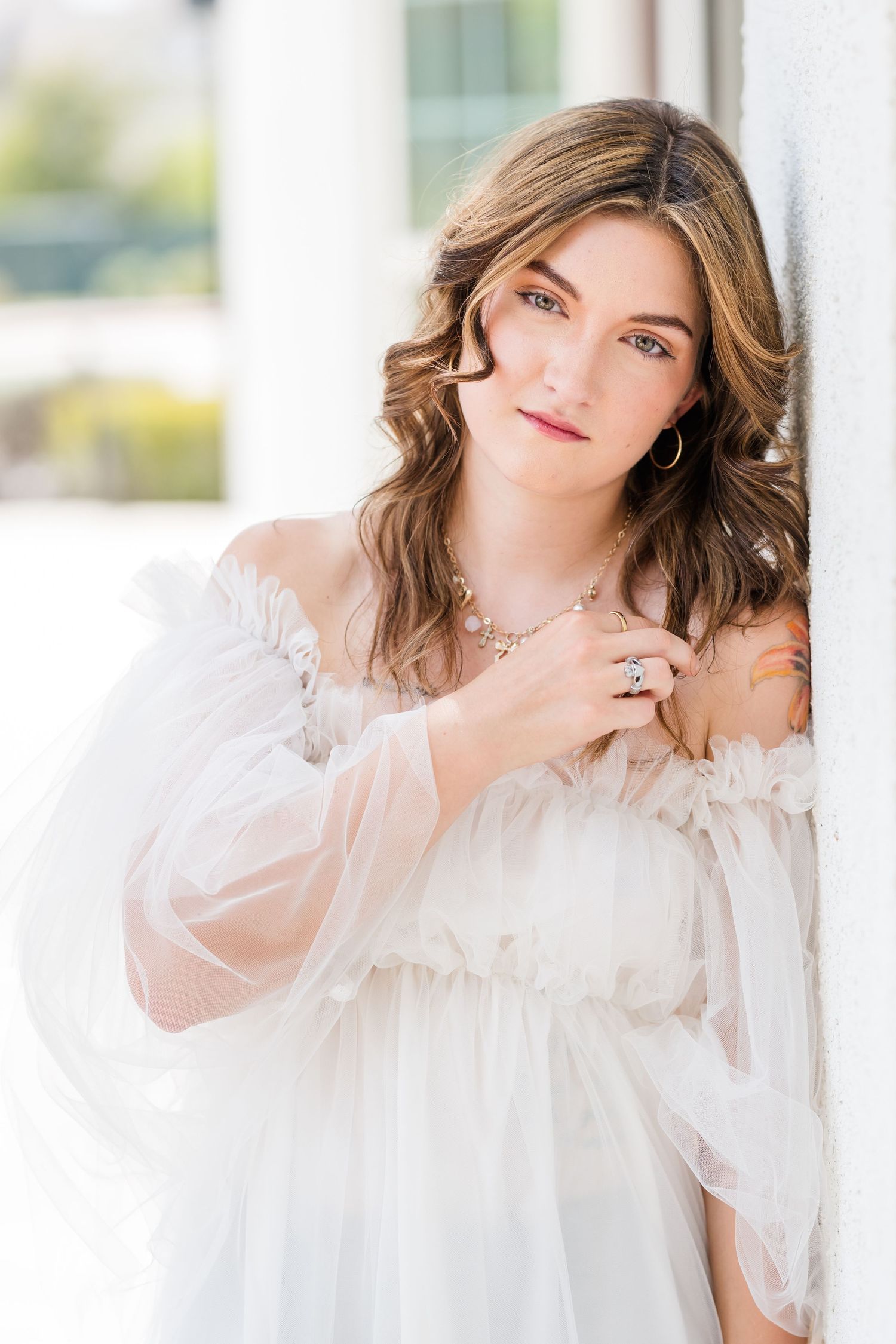 Young woman in white off-shoulder sheer dress with jewelry, leaning against white column.