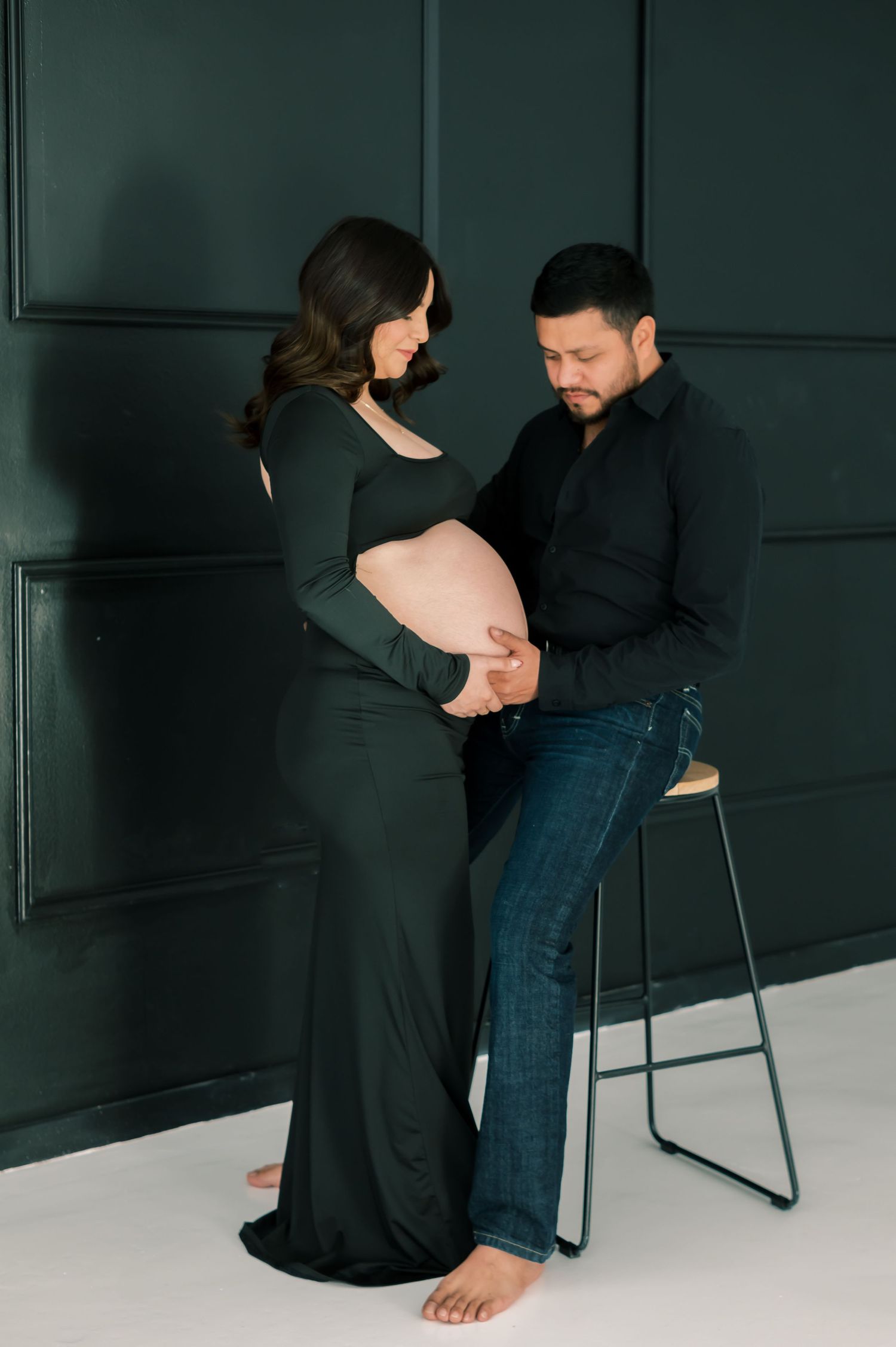 Expecting couple in dark green outfits tenderly cradling baby bump during elegant maternity photo session in studio.