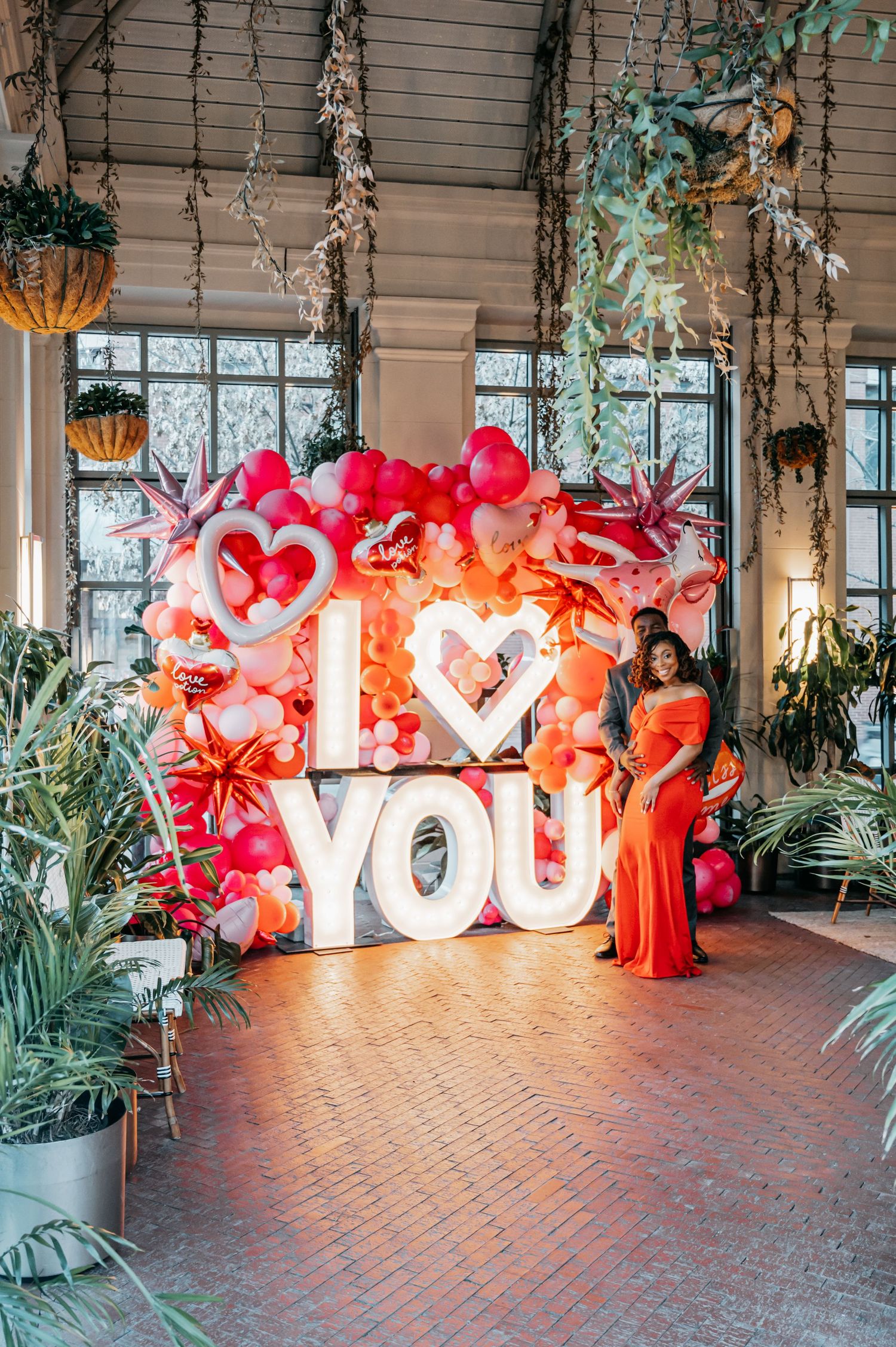 Couple poses in front of illuminated I Love You sign with red and pink balloon arch in decorated venue.