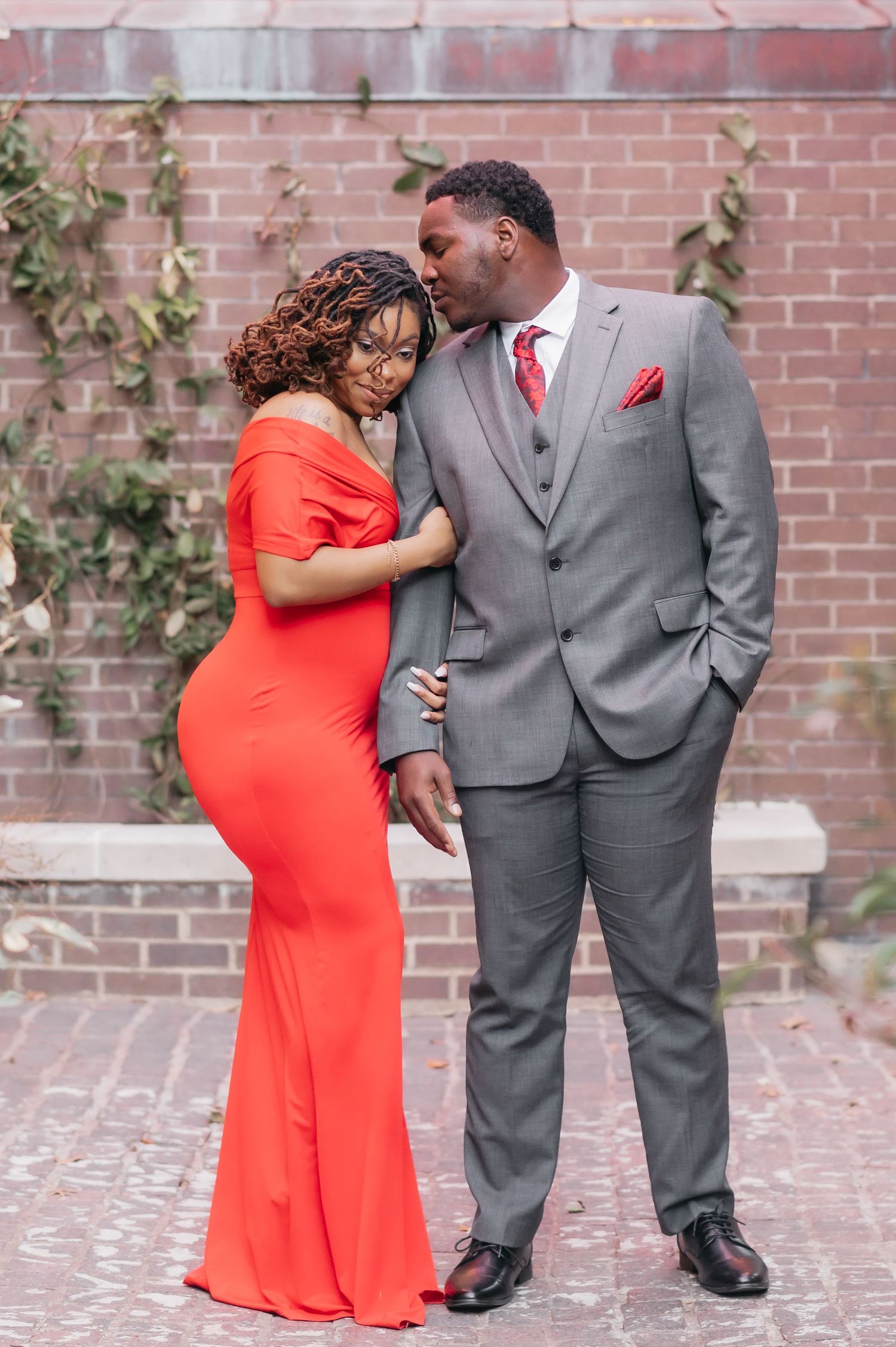 Couple in elegant red dress and gray suit sharing a romantic moment against a brick wall.