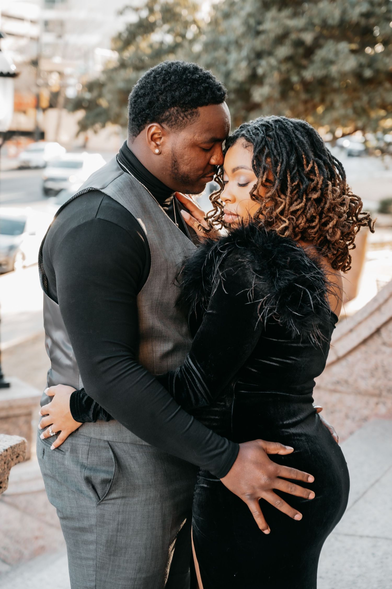 Couple sharing a romantic embrace outdoors, man in gray vest and woman in black feather-trimmed velvet dress.