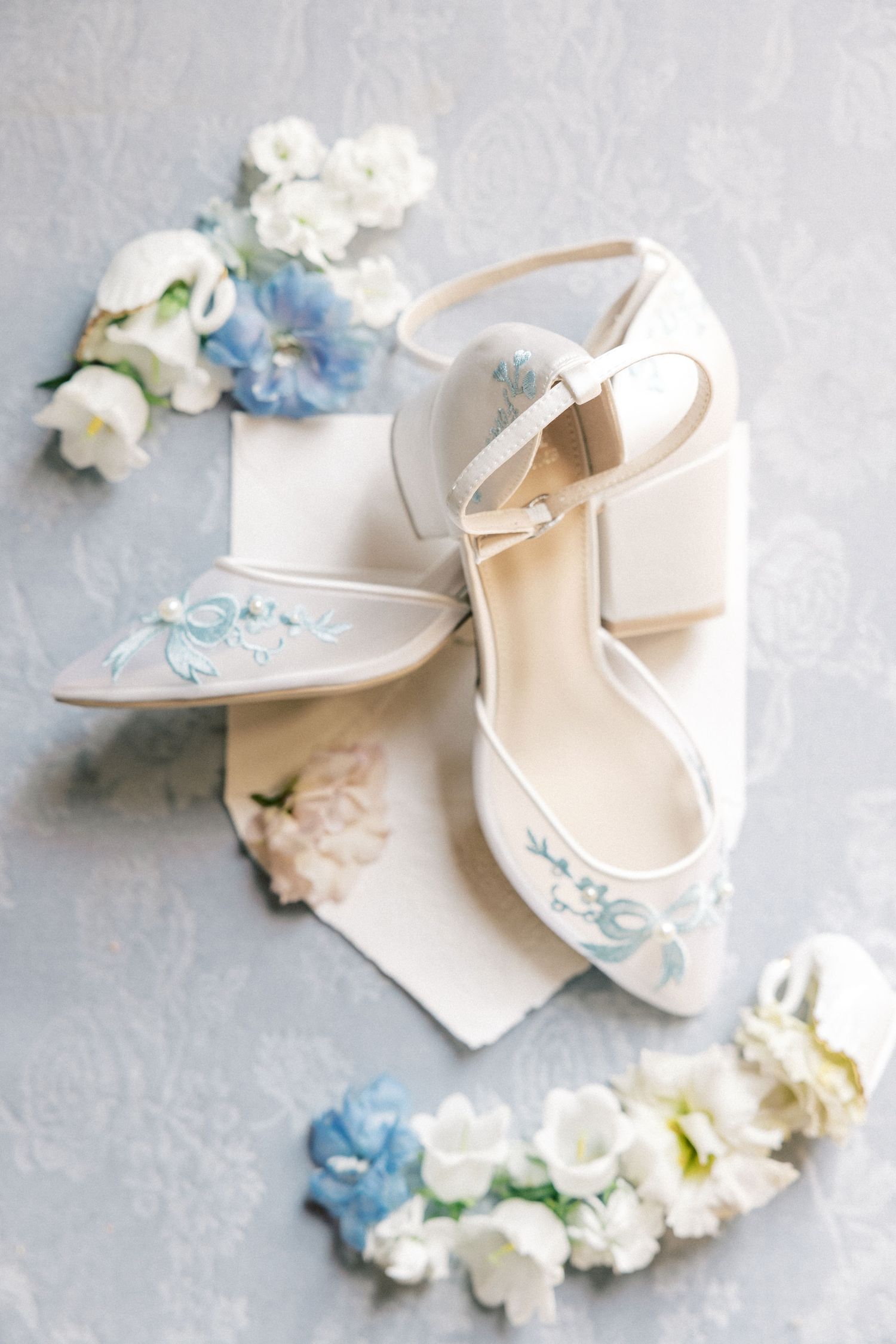 Elegant white bridal heels with blue floral embroidery and ankle straps, surrounded by delicate white and blue flowers.