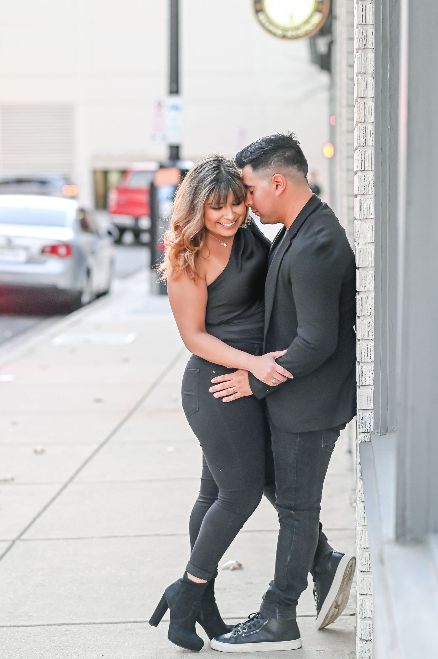 Couple in black outfits sharing a tender embrace while leaning against a brick building on a city street.