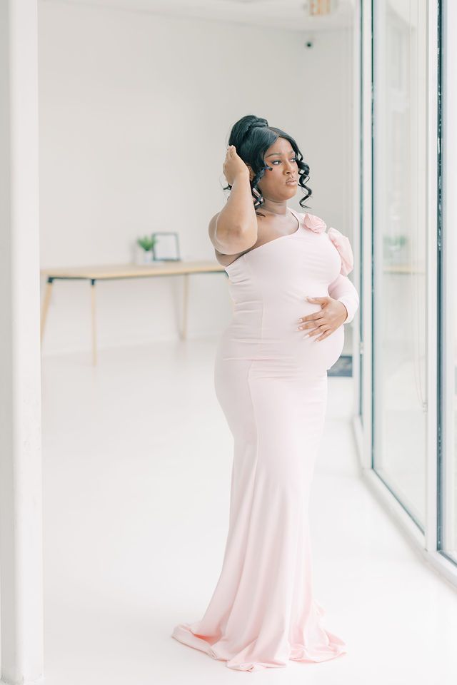 Pregnant woman in blush pink form-fitting gown posing near floor-to-ceiling windows.