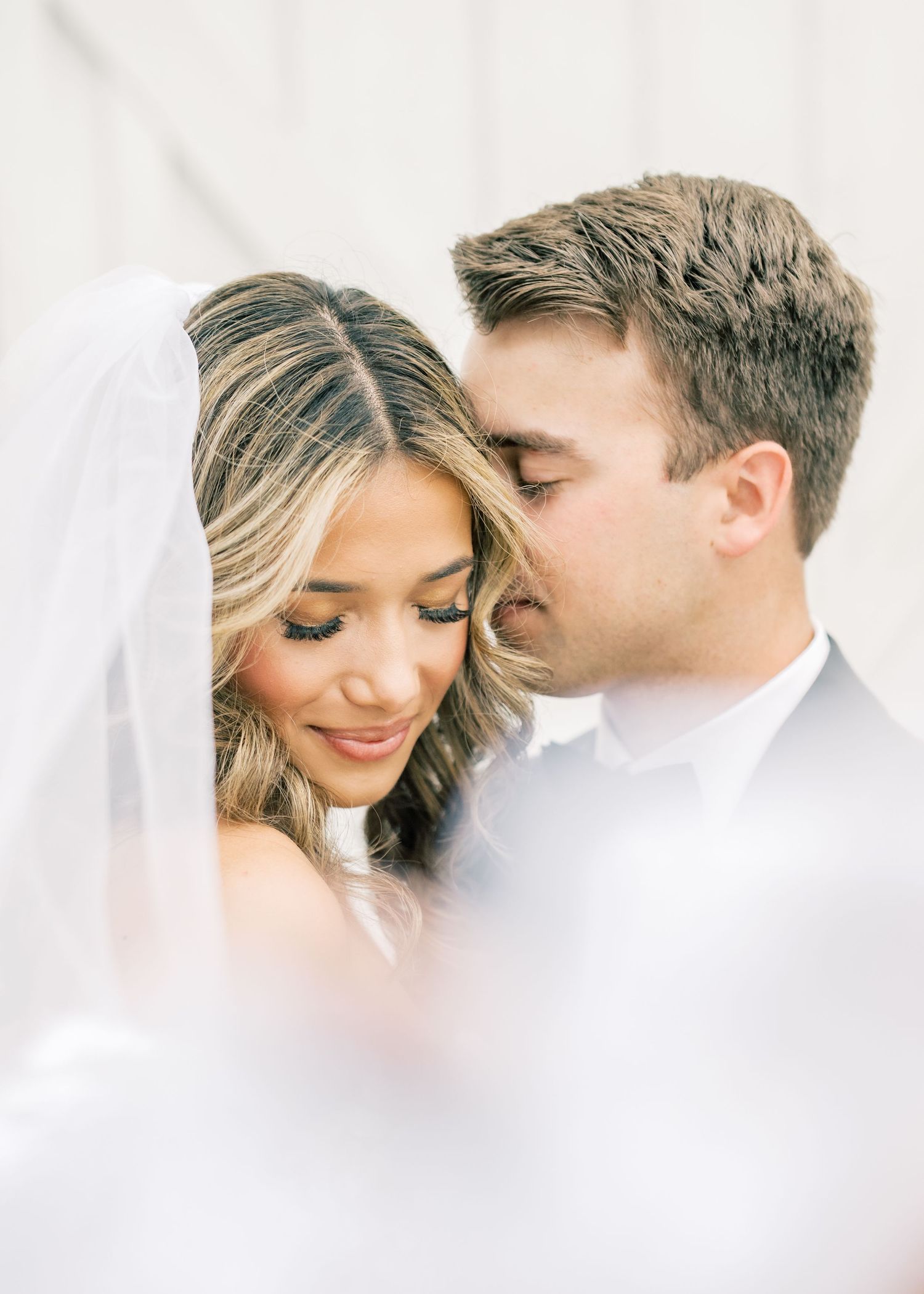 Romantic close-up of groom kissing brides cheek as she looks down smiling beneath her bridal veil.
