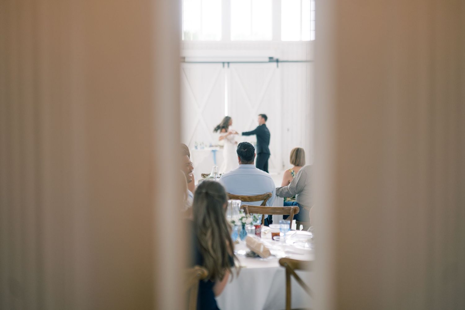 Bride and groom share their first dance viewed through a doorway as guests watch at reception tables.