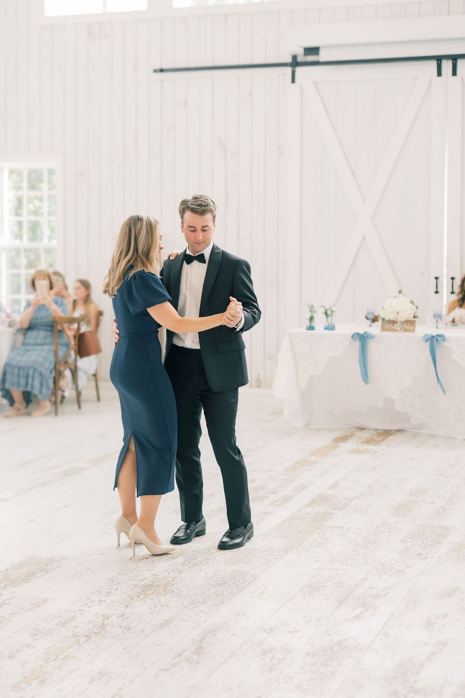 Groom dancing with mother in navy dress on white barn reception dance floor.