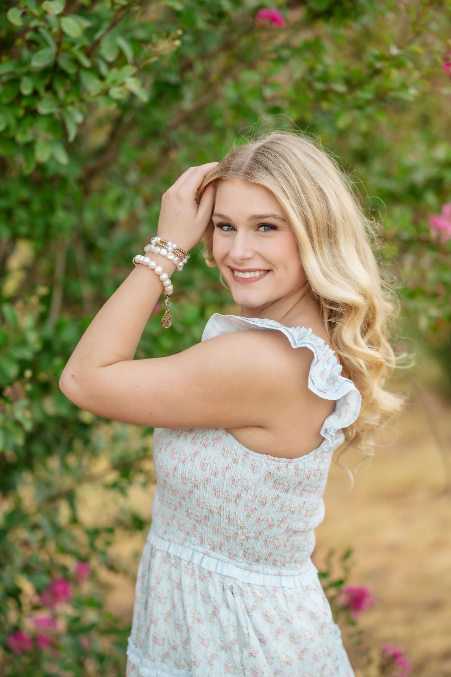 Smiling blonde woman in floral sundress with pearl bracelets posing outdoors among lush green bushes and pink flowers.