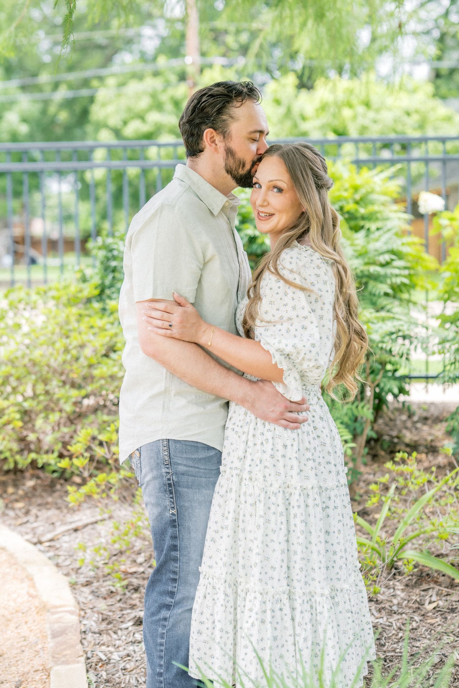 Man kissing womans head as she smiles in a sunlit garden setting.