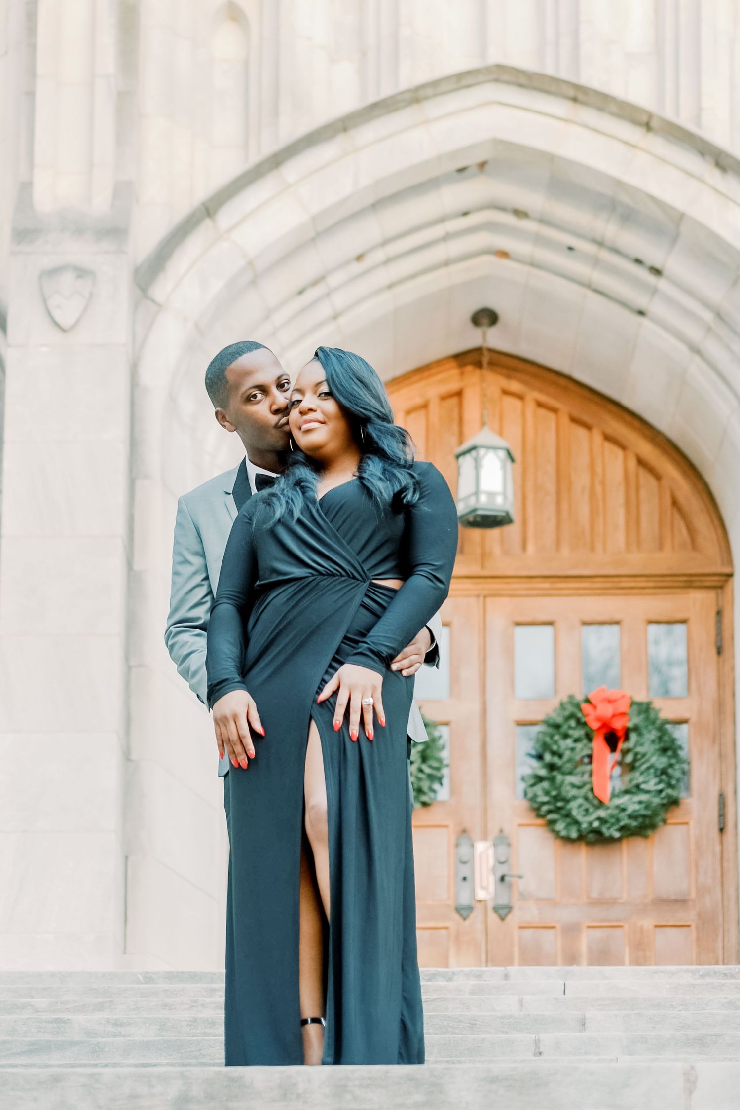 Couple embracing on church steps, man kissing woman's cheek, both dressed in elegant dark formal wear.
