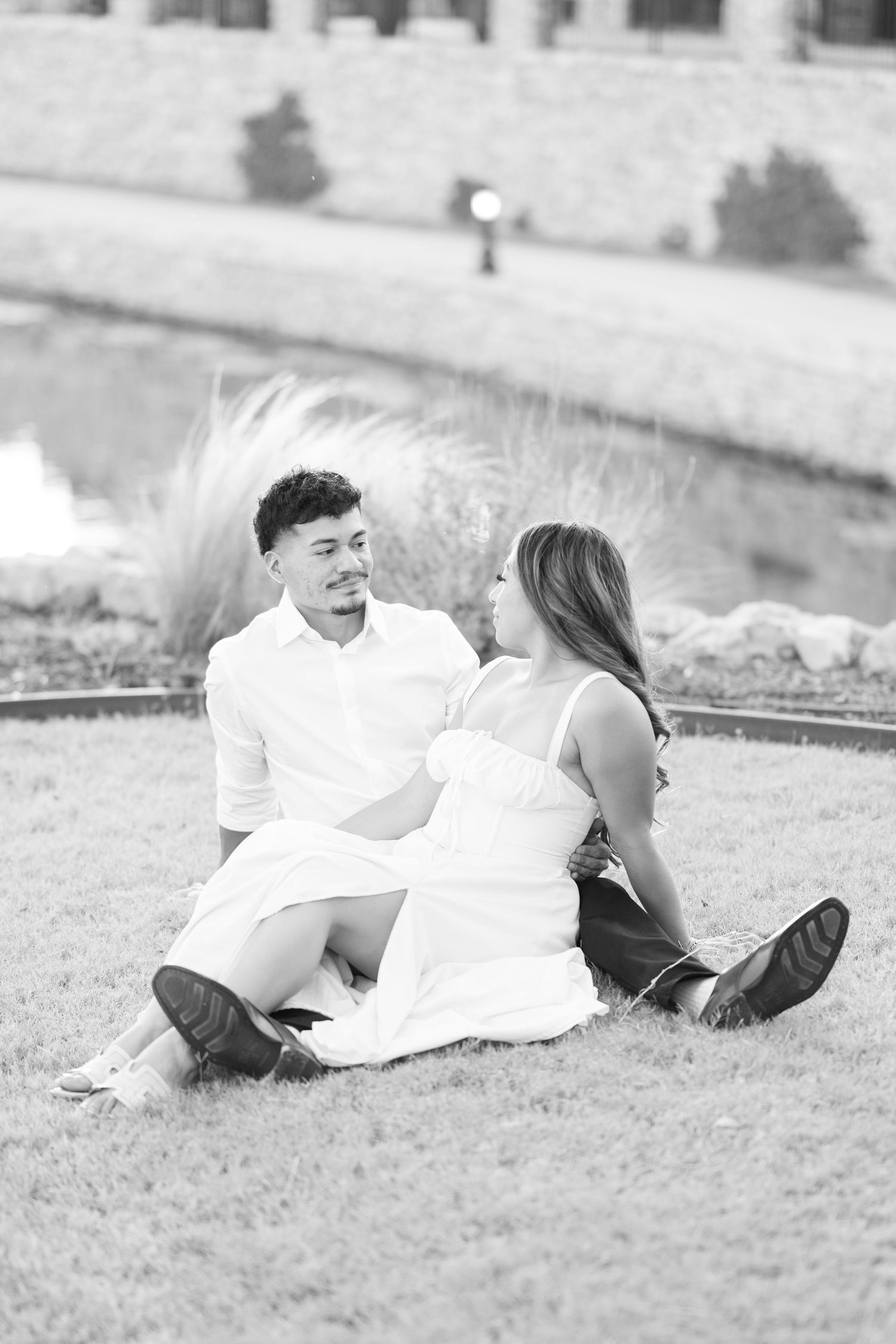Black and white photo of an engaged couple sitting together on grass, gazing at each other lovingly.
