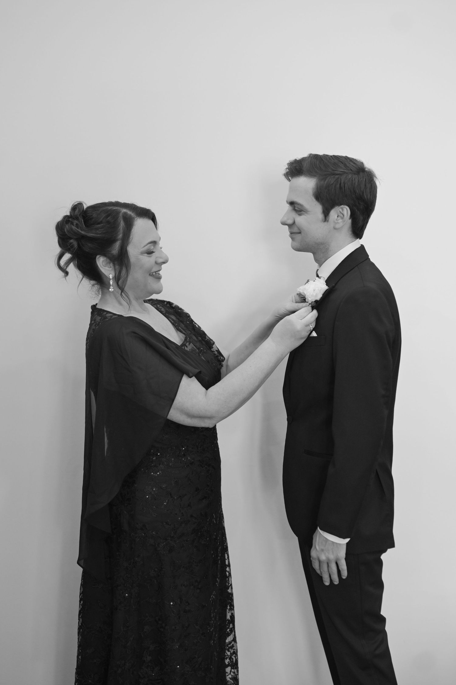 Black and white photo of mother pinning boutonniere on groom in black tuxedo before wedding ceremony.