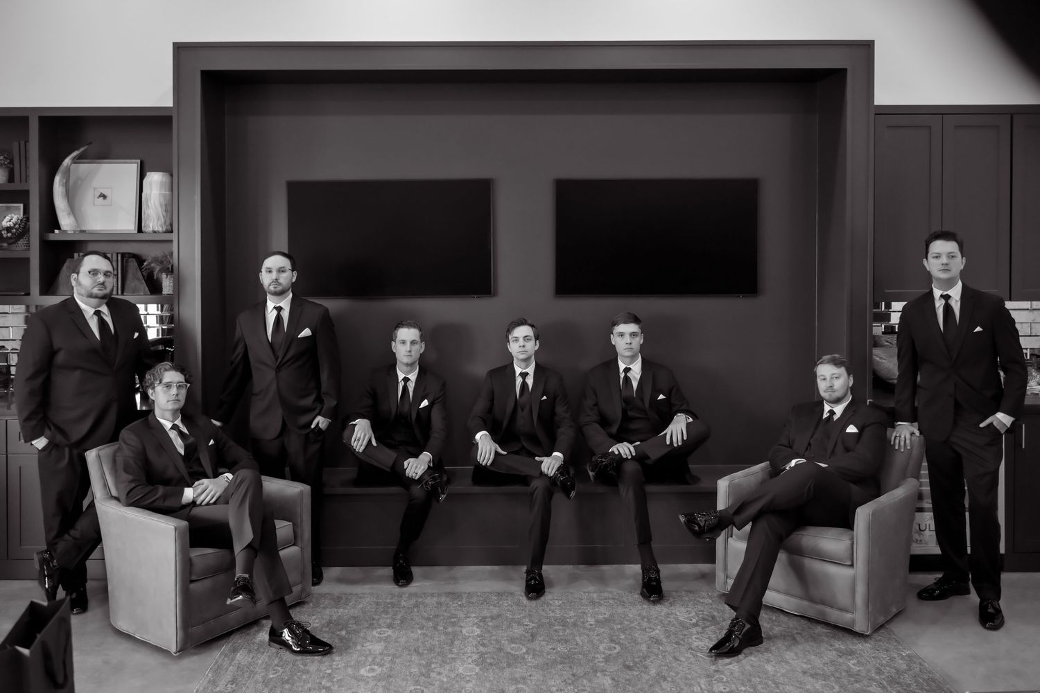 Black and white photo of nine men in elegant suits posing together in a modern lounge setting.