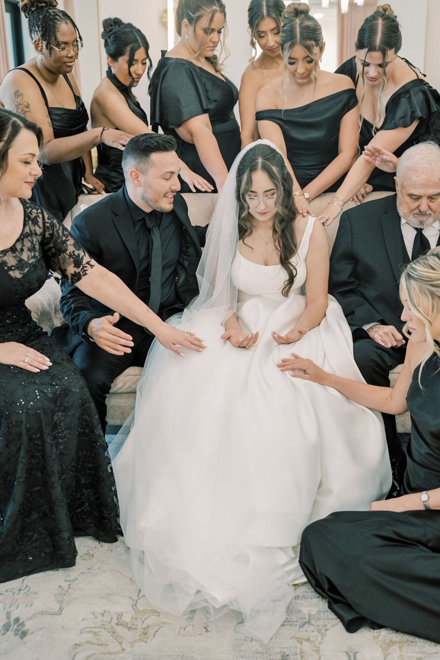 Bride in white gown surrounded by bridesmaids in black dresses during an intimate wedding prayer moment.