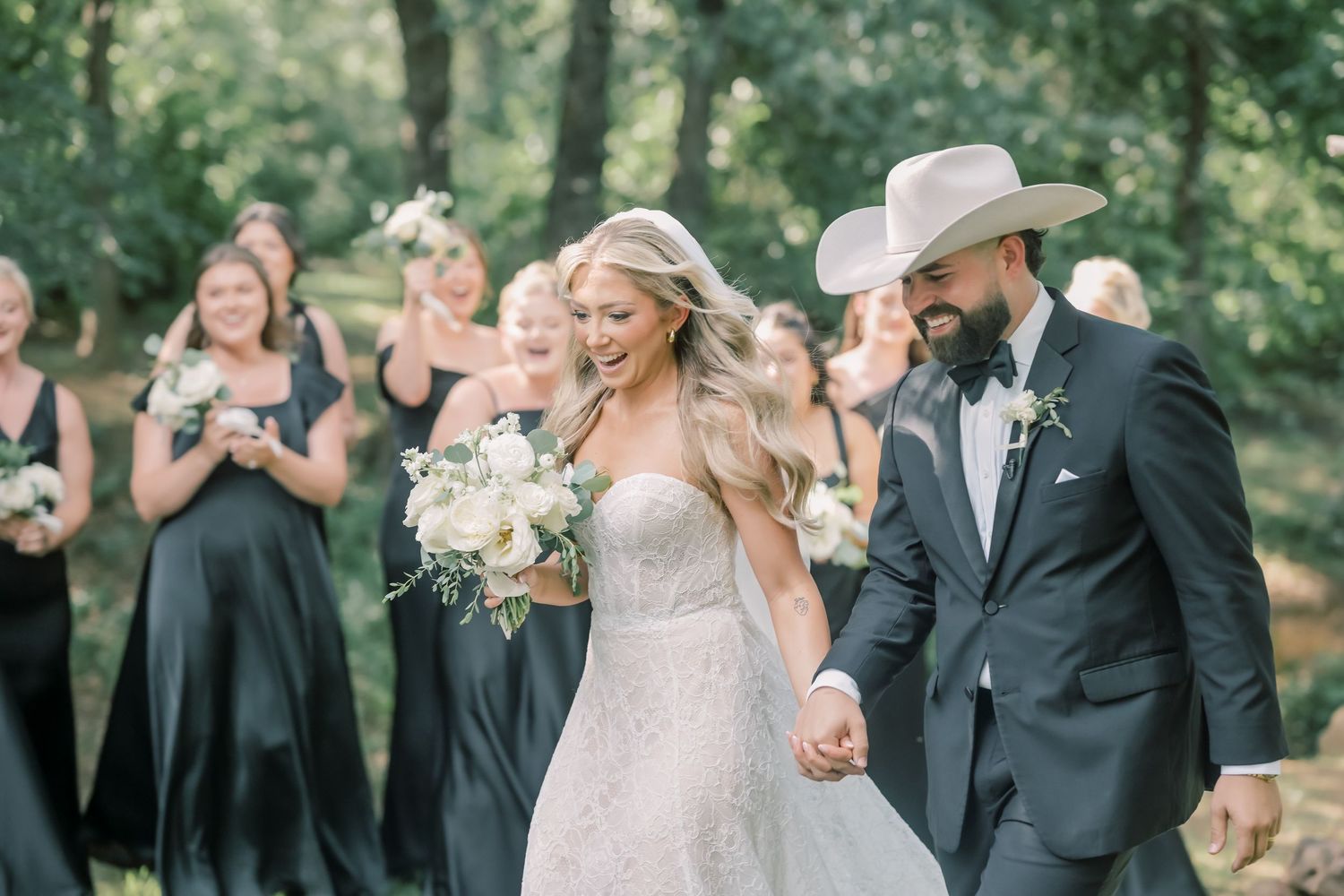 Joyful bride and groom in cowboy hat walk hand-in-hand with bridesmaids in black dresses at outdoor wedding.