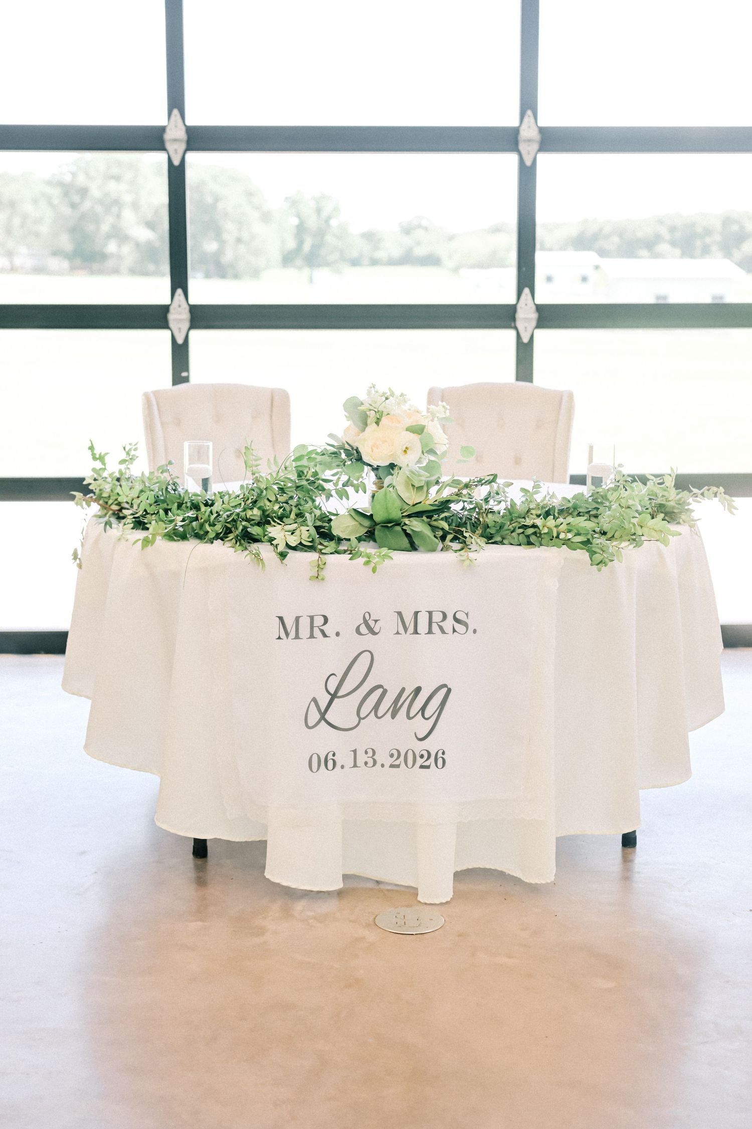 Sweetheart table with white tablecloth reading Mr. and Mrs. Lang 06.13.2026 with floral garland.