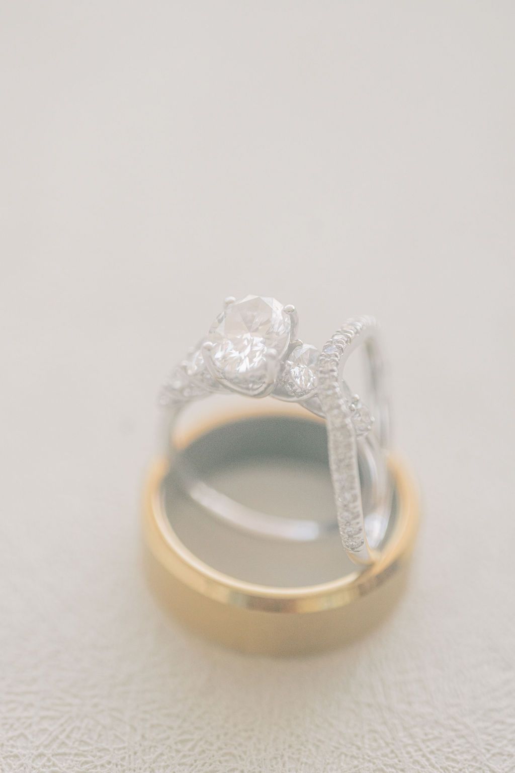 Diamond engagement ring with oval stone resting on gold wedding band displayed on white surface.